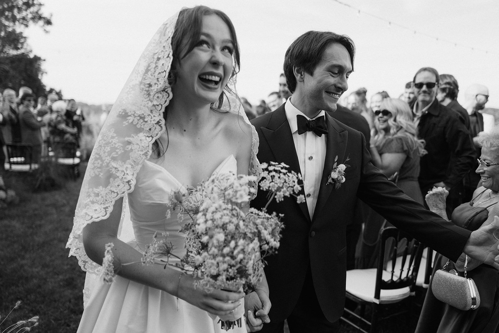 A black and white photo of a newlywed couple at their outdoor wedding, the bride wearing a lace veil and holding a bouquet, the groom in a tuxedo with a boutonniere, surrounded by smiling guests.