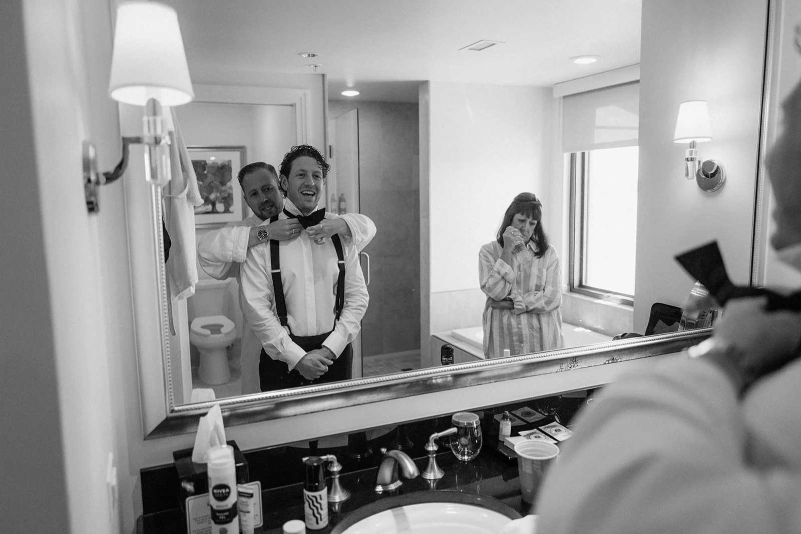 A man in tuxedo and suspenders stands in front of a mirror as another man helps him with his bow tie. A woman to the right looks on thoughtfully. The bathroom counter has various toiletries.
