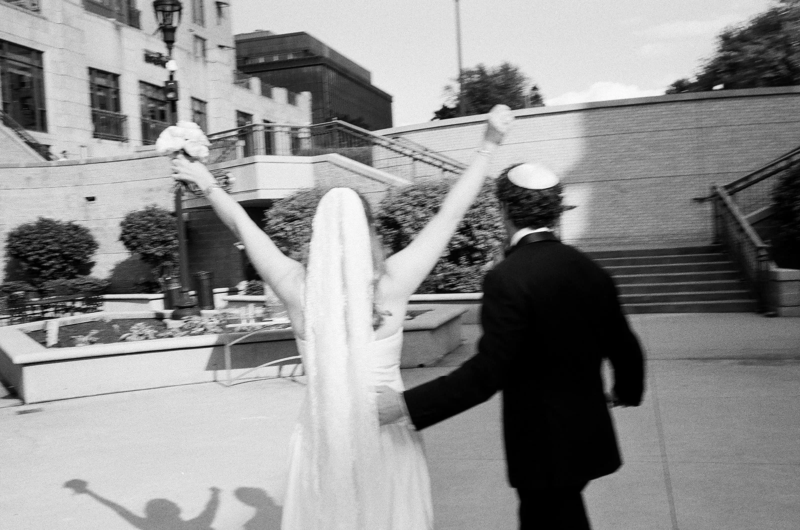 A bride and groom at a wedding ceremony outside in a city, with the bride raising her arms and holding a bouquet, and the groom holding her dress from behind. The photo is in black and white.