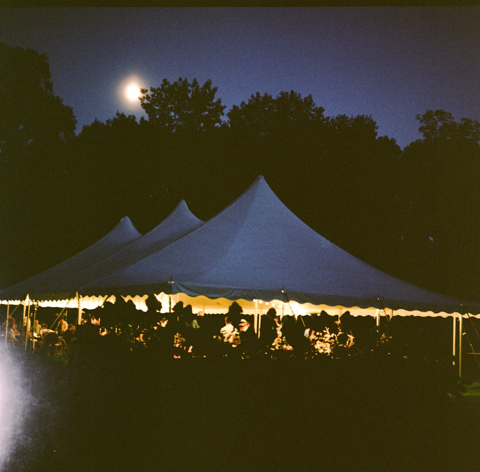 Nighttime outdoor gathering under a large illuminated white tent with people inside, set against a dark sky with a bright moon and trees in the background. shot on film on the rolleiflex in a documentary style. 