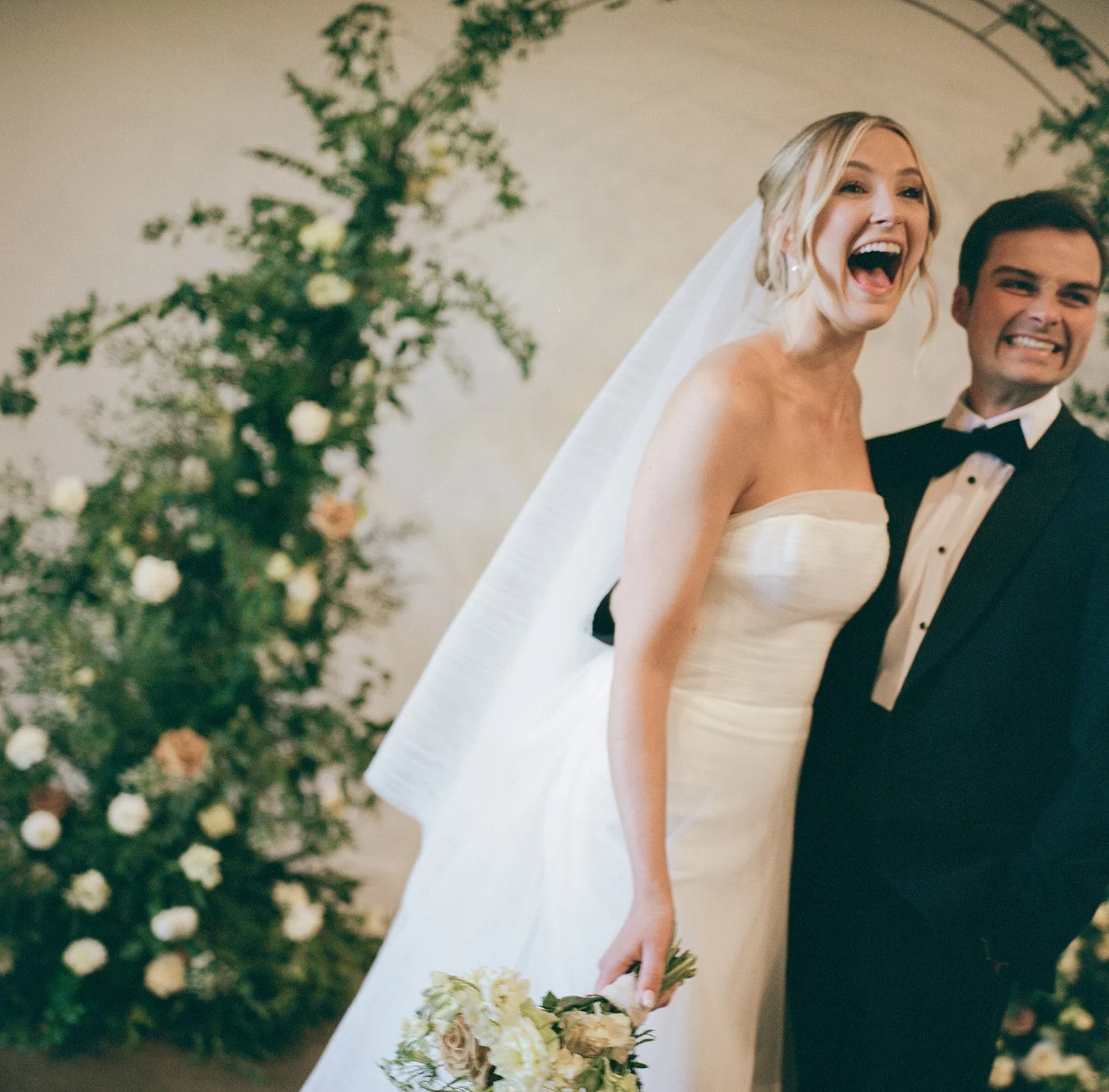 A newlywed couple is smiling and posing for a photo indoors, standing in front of a floral arch with greenery and white flowers. The bride is wearing a strapless white wedding gown and holding a bouquet of flowers, while the groom is in a black tuxed