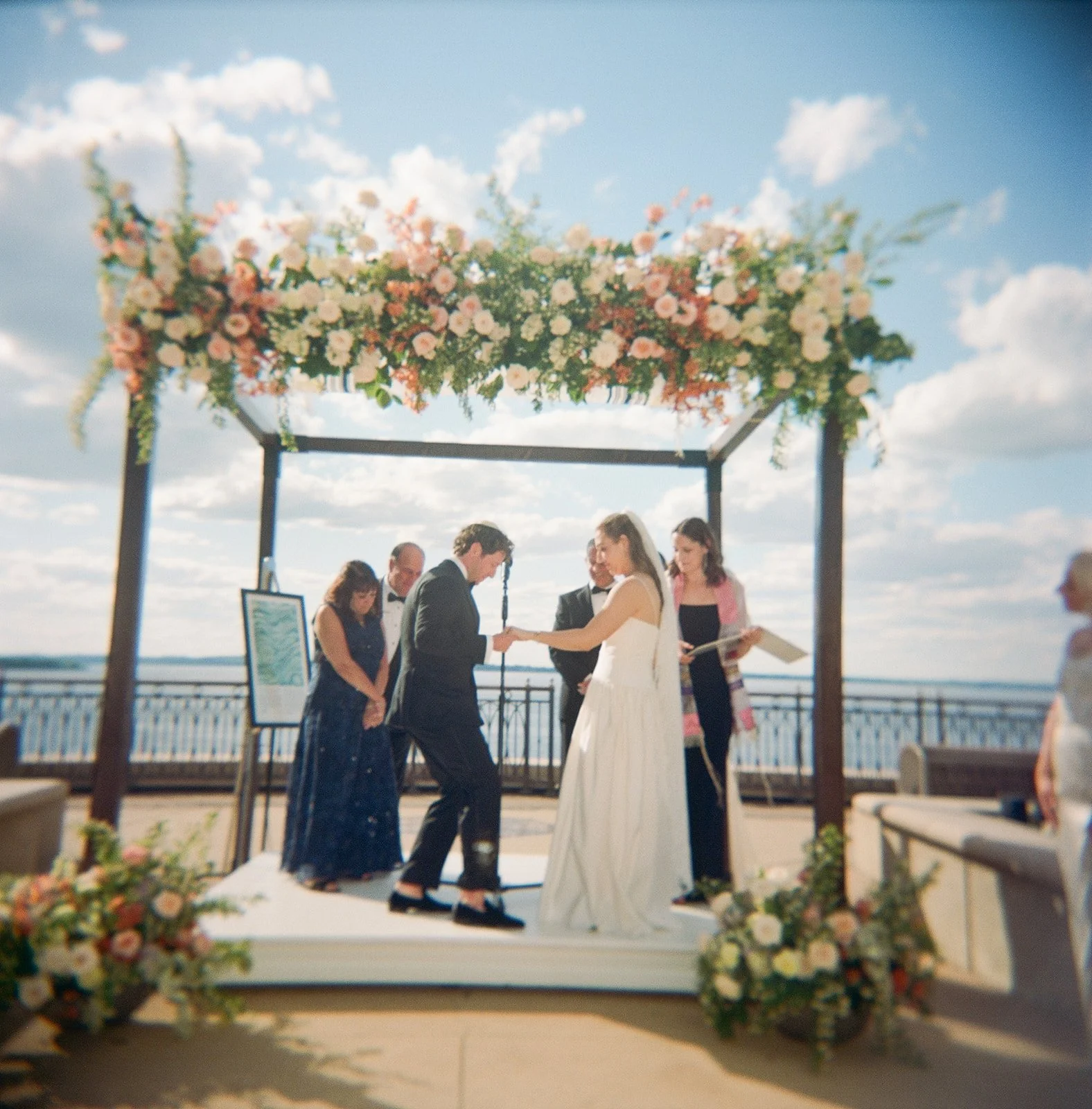 A wedding ceremony taking place outdoors under a floral canopy with a blue sky and clouds in the background. The bride and groom are exchanging vows on a small white platform, surrounded by guests and floral arrangements.