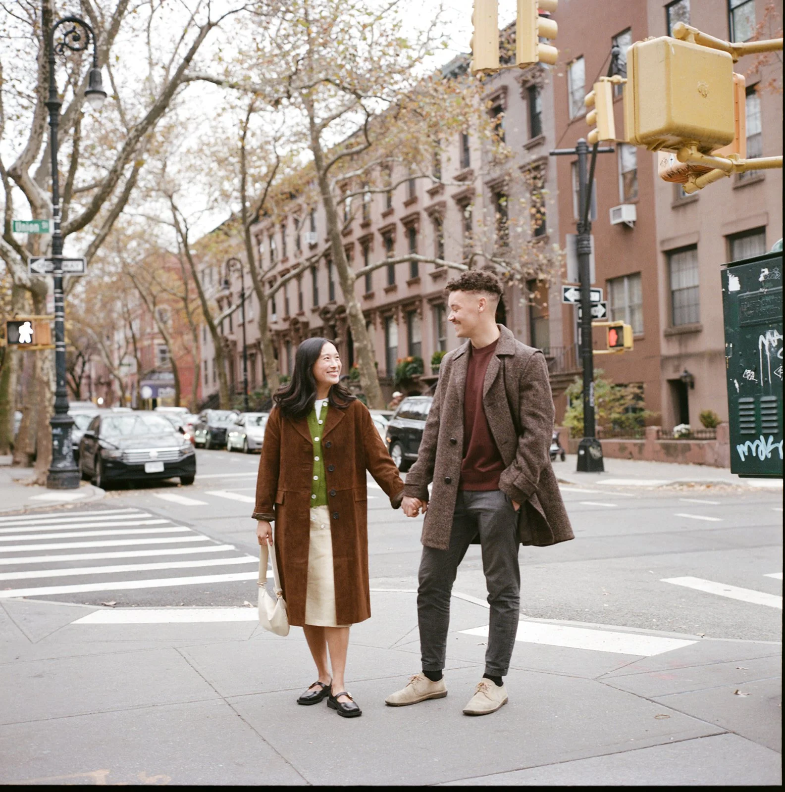 A couple holding hands and walking across a city street corner with brownstone buildings and trees with fall foliage, smiling at each other in cobble hill, brooklyn.