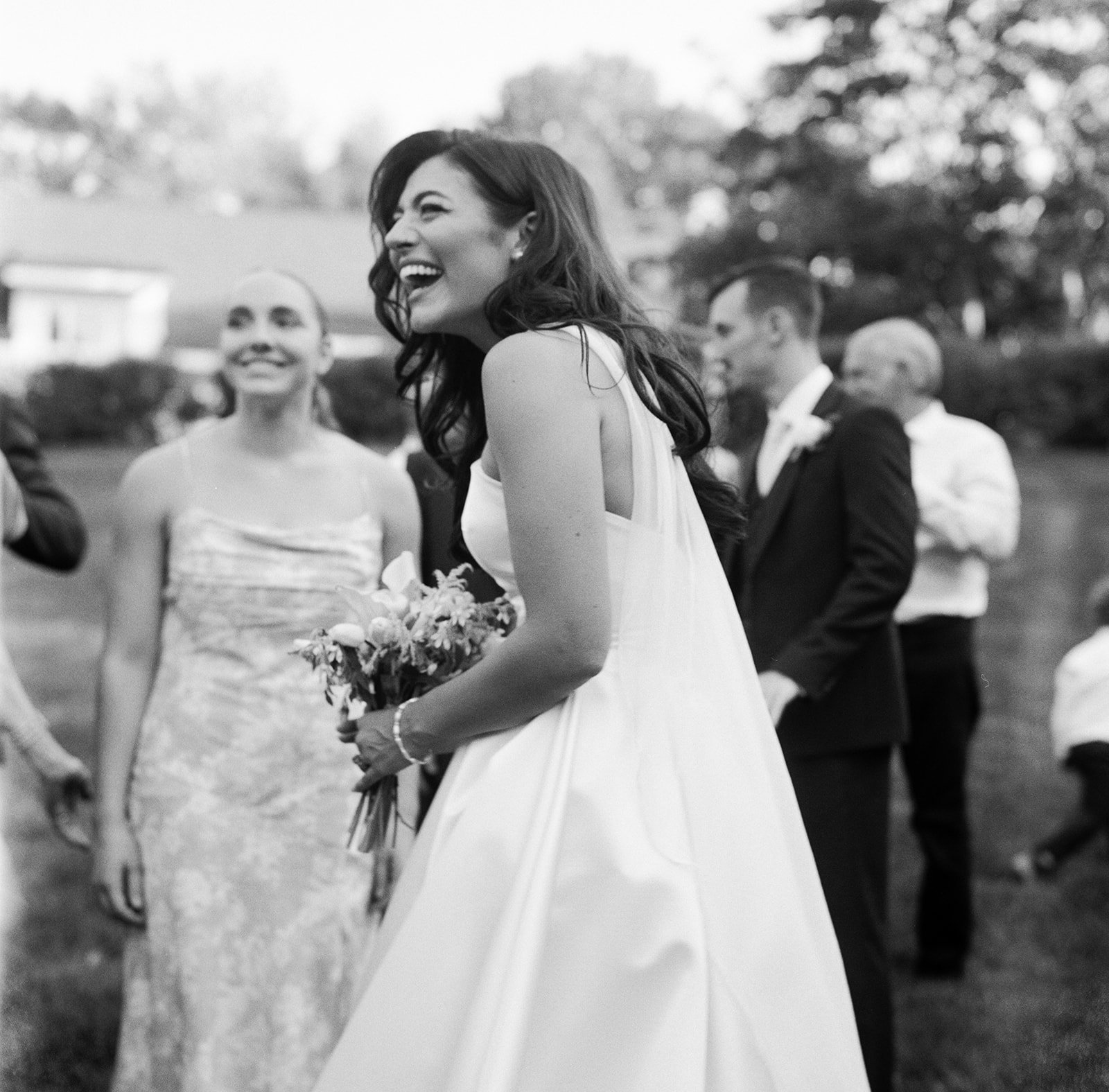 Black and white photo of a happy bride holding a bouquet at a wedding reception outdoors, surrounded by guests in formal attire, with trees in the background. shot on film on the rolleiflex in a documentary style. 