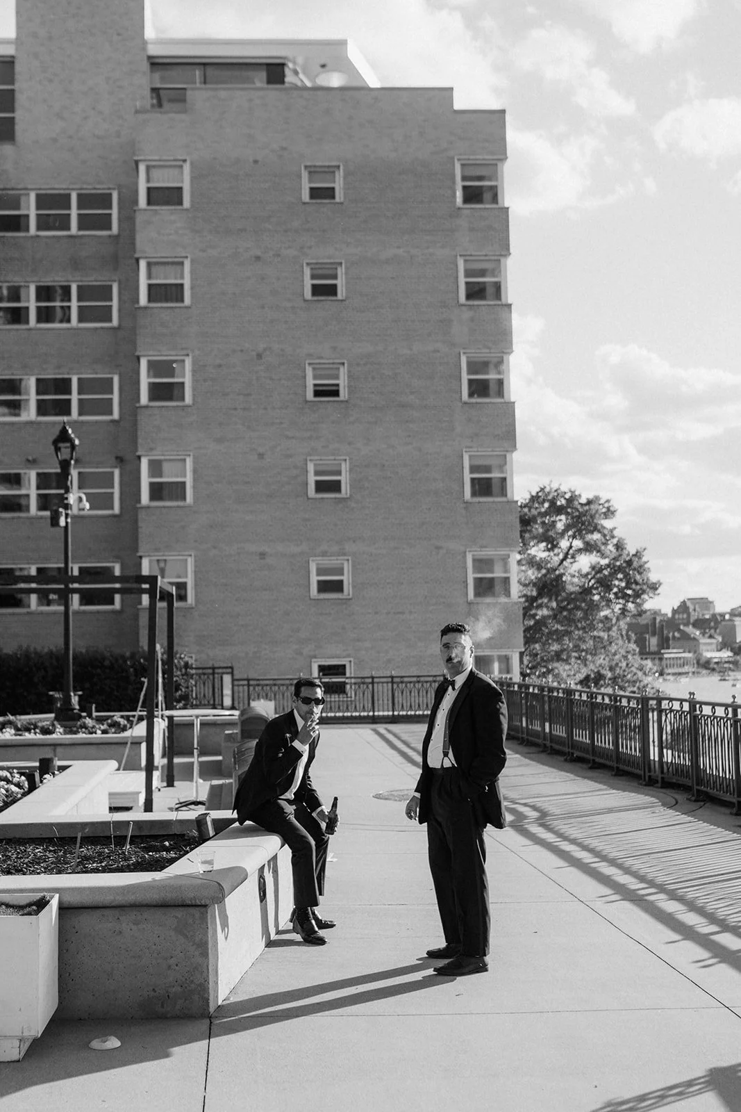 Two men in tuxedos standing and sitting outside on a terrace, with a tall building and cloudy sky in the background.