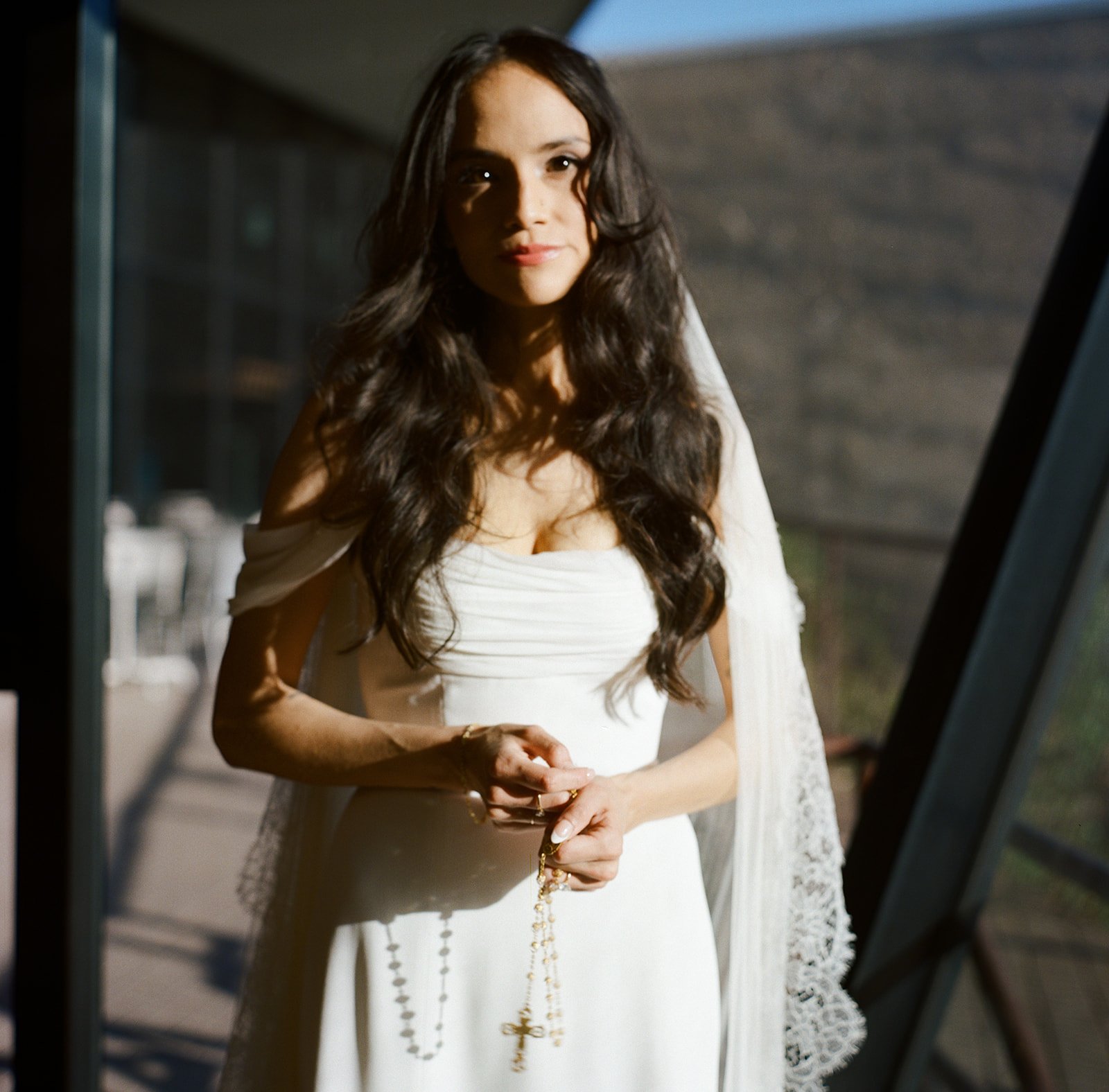 A woman in a white dress standing near a glass window, holding a rosary with a cross in her hands.