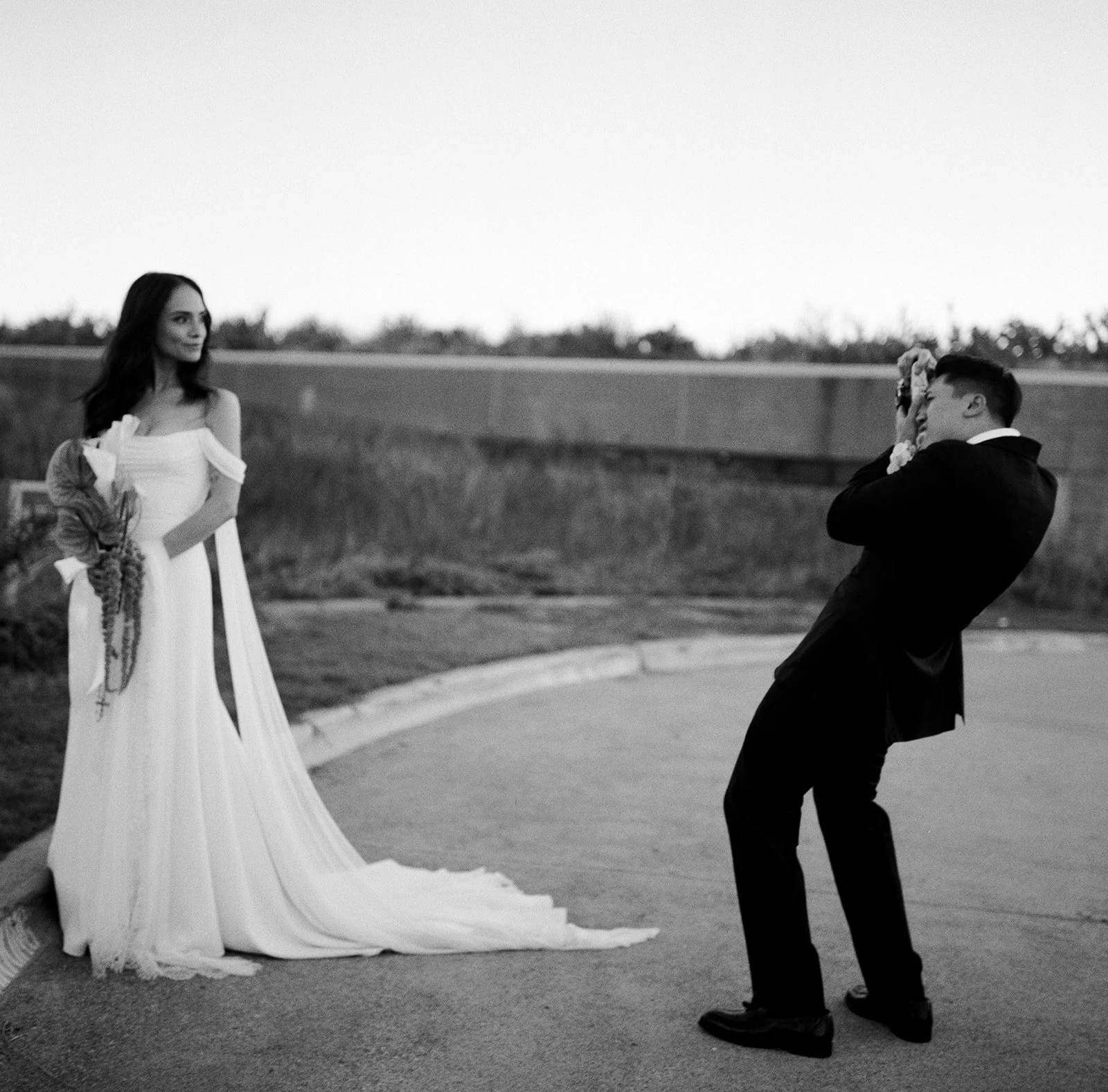 A bride in a white wedding dress holding a bouquet, standing on a curb, as a groom in a tuxedo kneels and takes a photo of her outdoors during sunset.