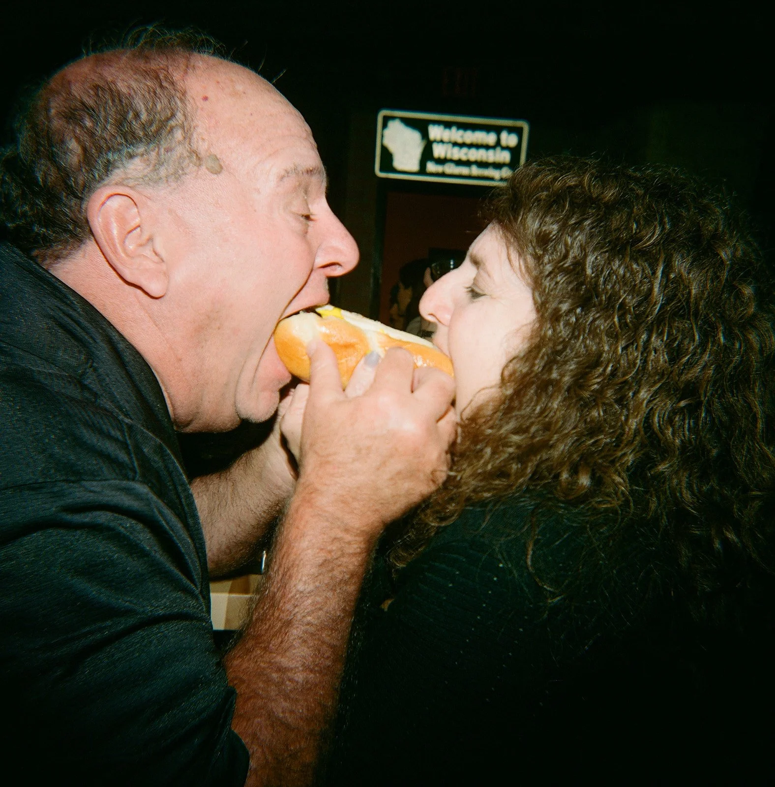 A man and woman sharing a hot dog, facing each other with the hot dog between their mouths.