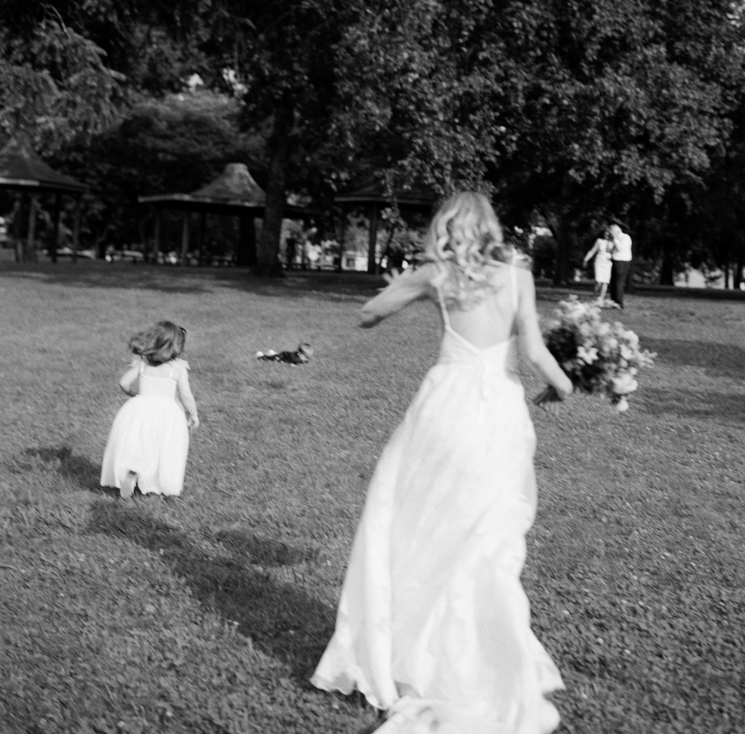 A bride in a wedding gown with a bouquet walking in a park with two children, a little girl in a white dress and a young girl in a white dress, and a couple dancing in the background under large trees.