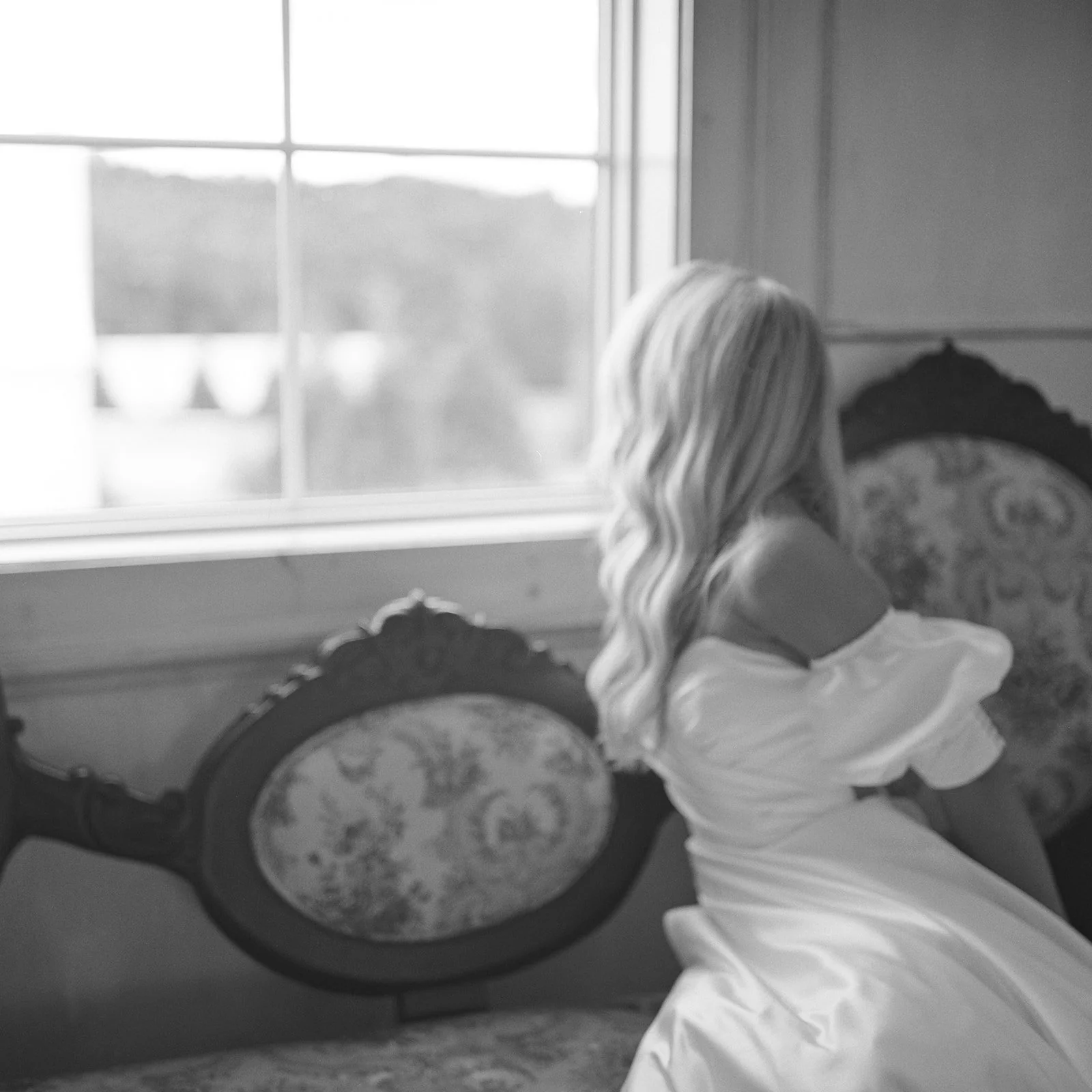 A woman with long, wavy blonde hair sitting on an ornate vintage sofa with floral upholstery, looking out a window.
