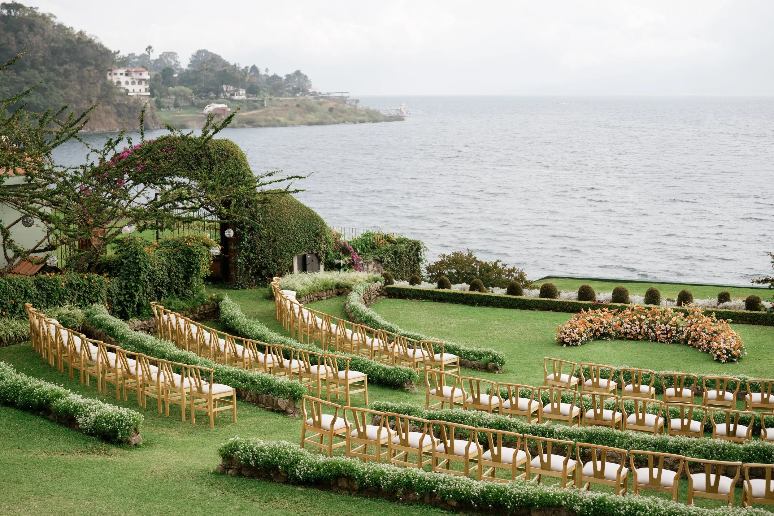 Outdoor wedding ceremony setup on a grassy lawn overlooking the ocean, with rows of wooden chairs and floral decorations.