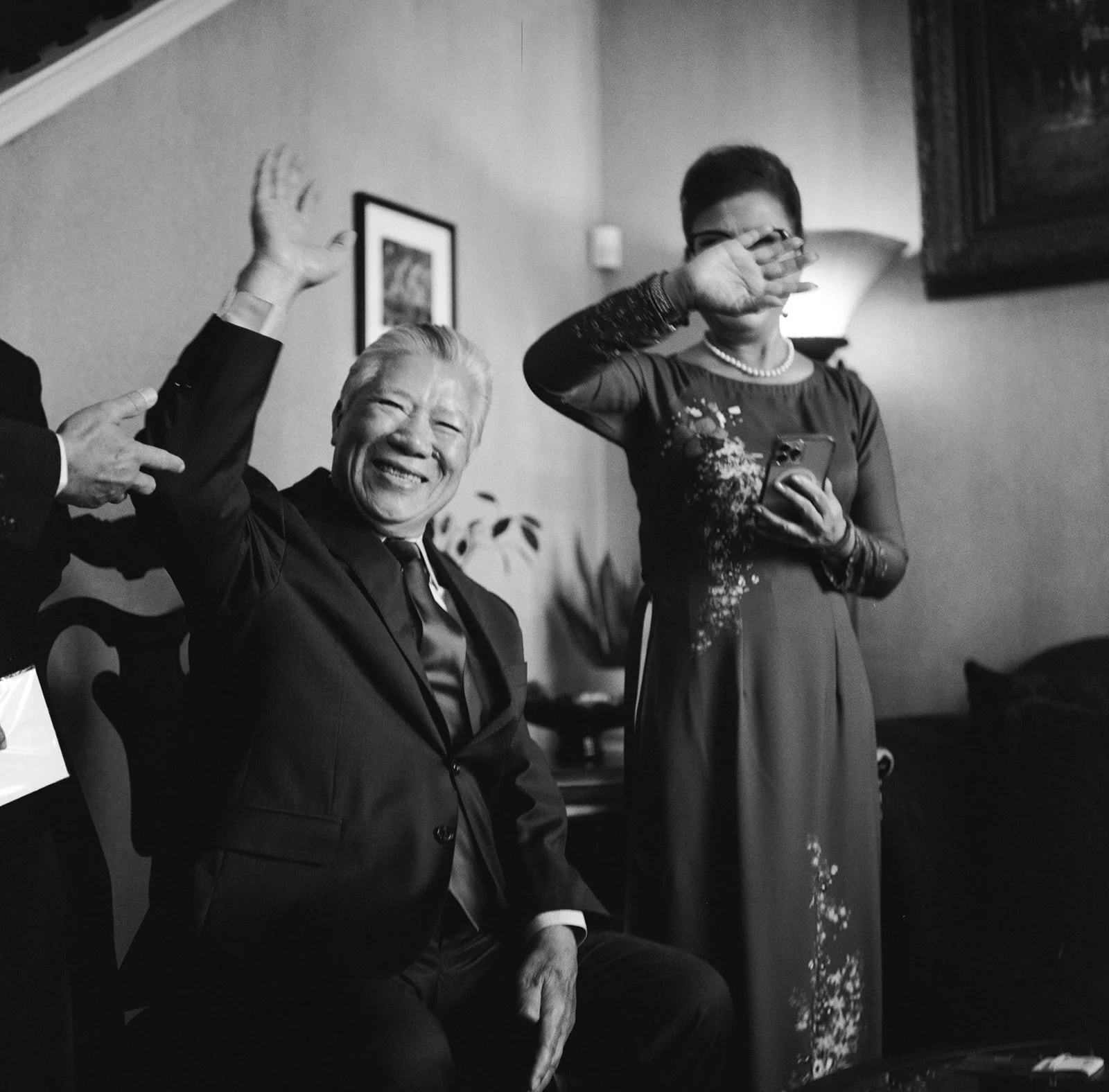A black and white photo of an elderly man in a suit and tie smiling and waving, while a woman standing next to him, holding a phone and wiping her eye, also smiling and waving.