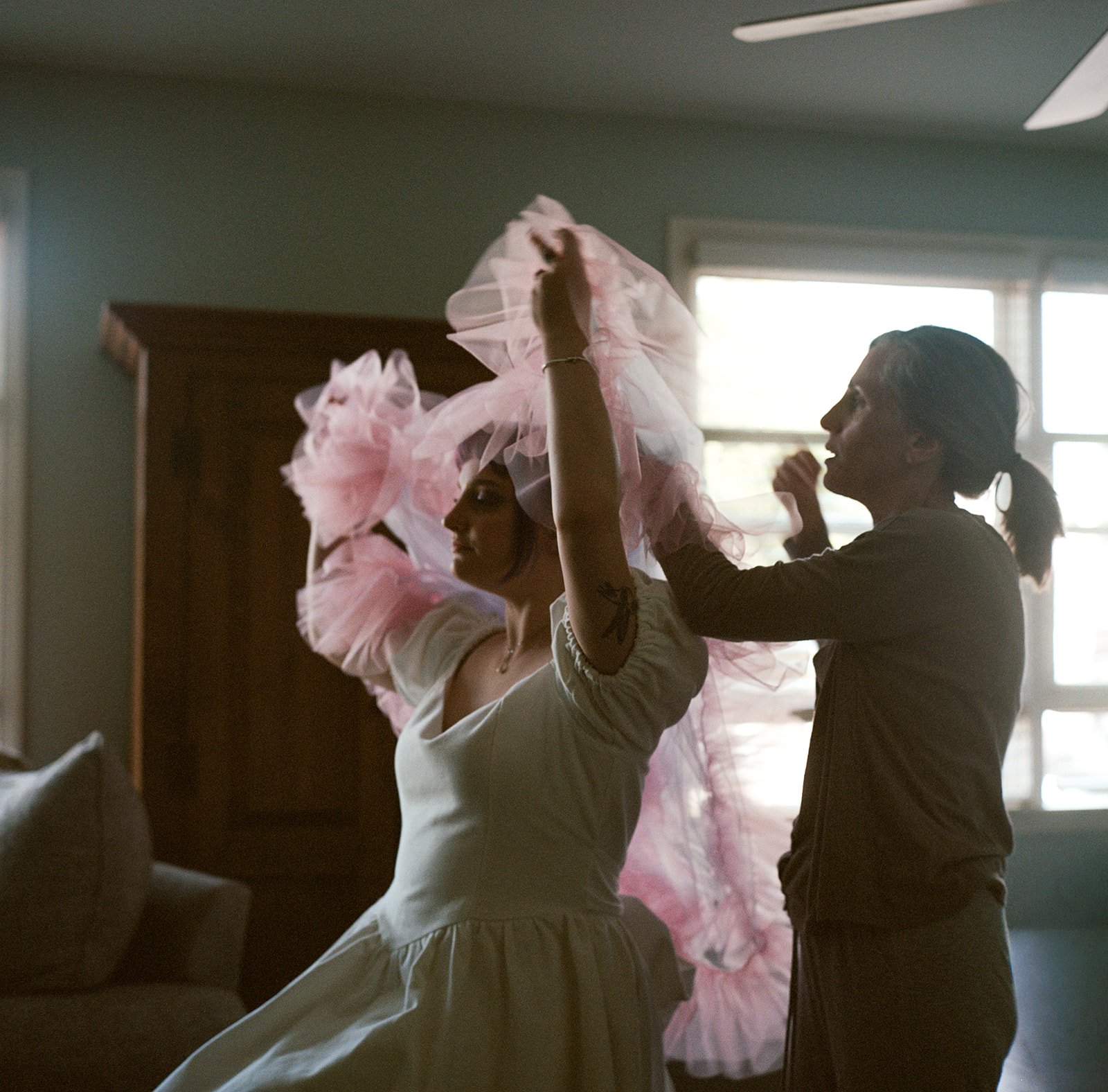 A woman in a light-colored dress is having pink tulle fabric placed over her shoulders by another woman in a darker top. They are inside a room with natural light coming through windows.
