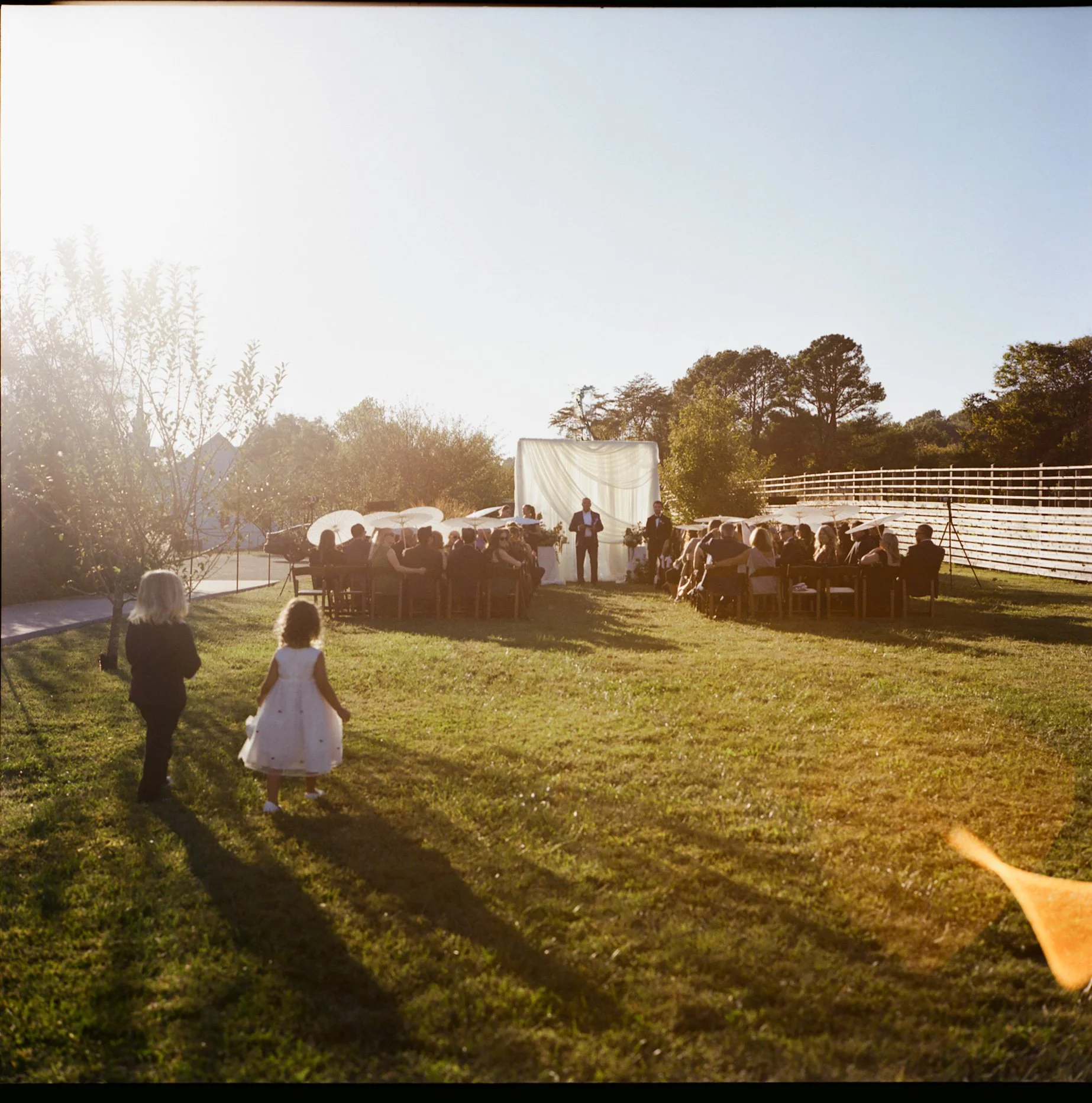 Outdoor wedding ceremony with guests seated at tables, two children walking on the grass in the foreground, sunset lighting, trees, and a white backdrop for the ceremony. shot on film on the rolleiflex in a documentary style. 
