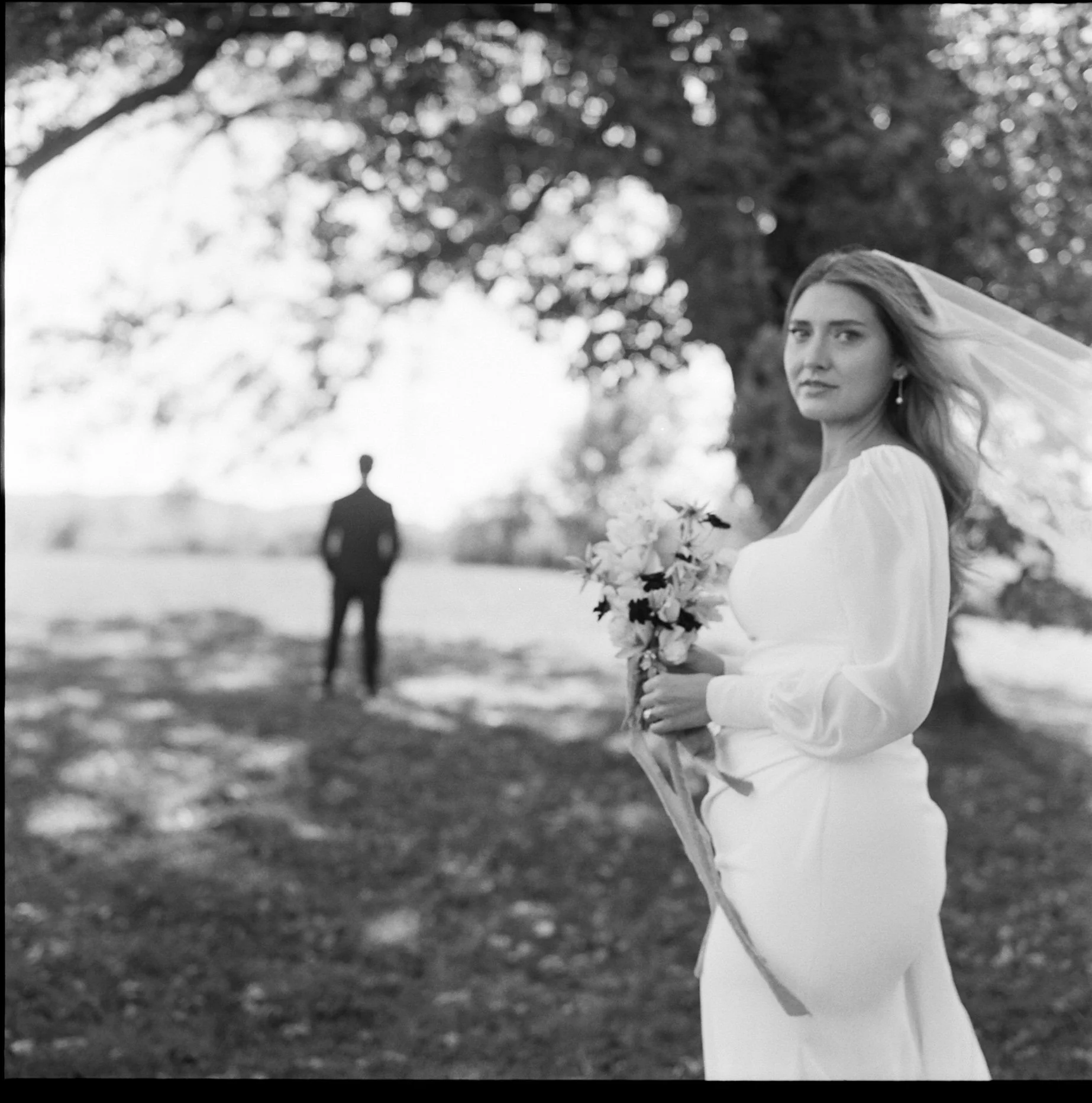 A woman in a white dress holding a bouquet of flowers outdoors, with a man in a suit standing in the background near a large tree. shot on film on the rolleiflex in a documentary style. 