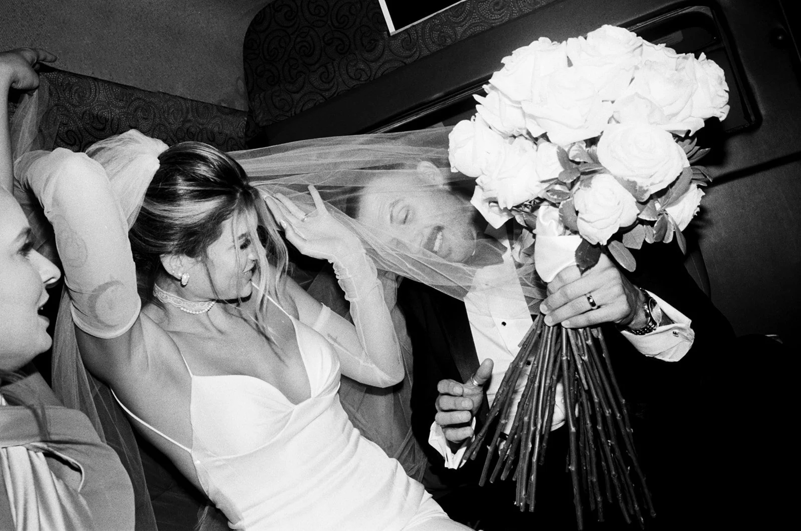 Bride in a wedding dress lying down, smiling, with a veil, surrounded by people, holding a bouquet of white roses.