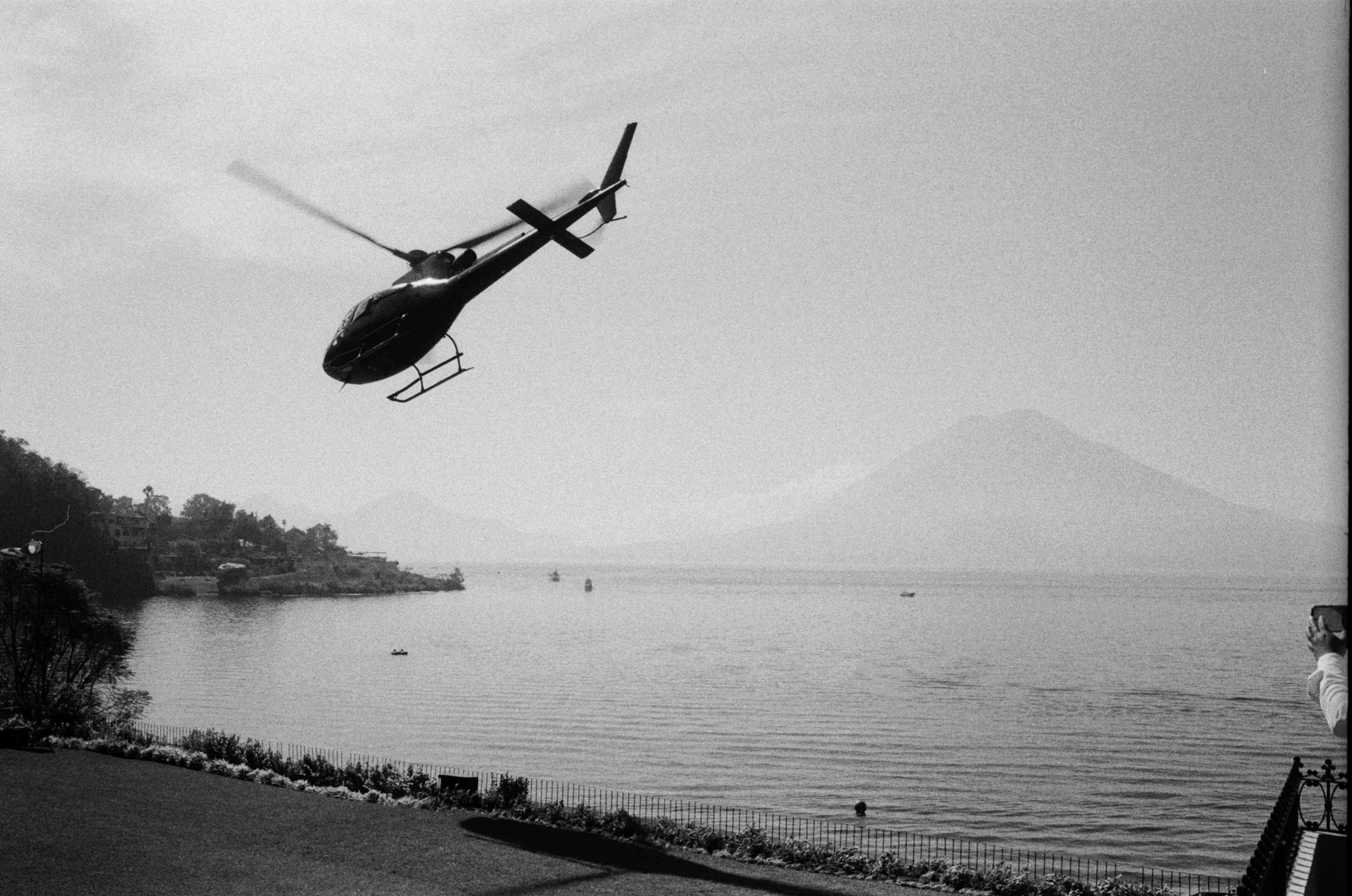 A black helicopter flying over a body of water with mountains in the background, a person taking a photo on the right side, and houses on the left side of the shoreline.