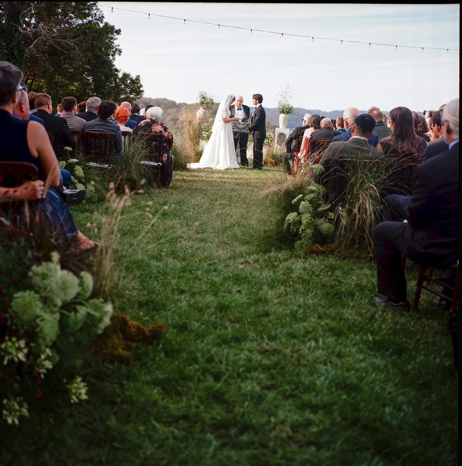 A wedding ceremony outdoors with the bride and groom standing before an officiant, surrounded by seated guests on both sides, with the setting featuring greenery and string lights overhead.
