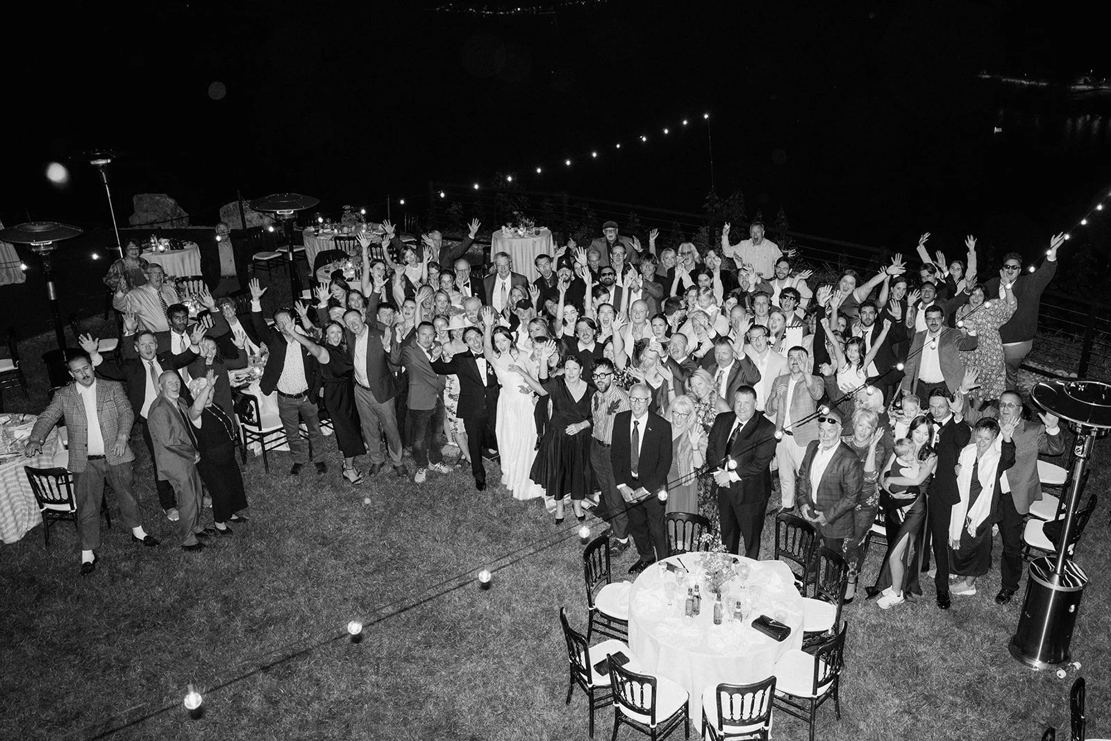 Black and white photo of a large group of people at a nighttime celebration outdoors, standing and raising their hands, with decorated tables and string lights in the background.