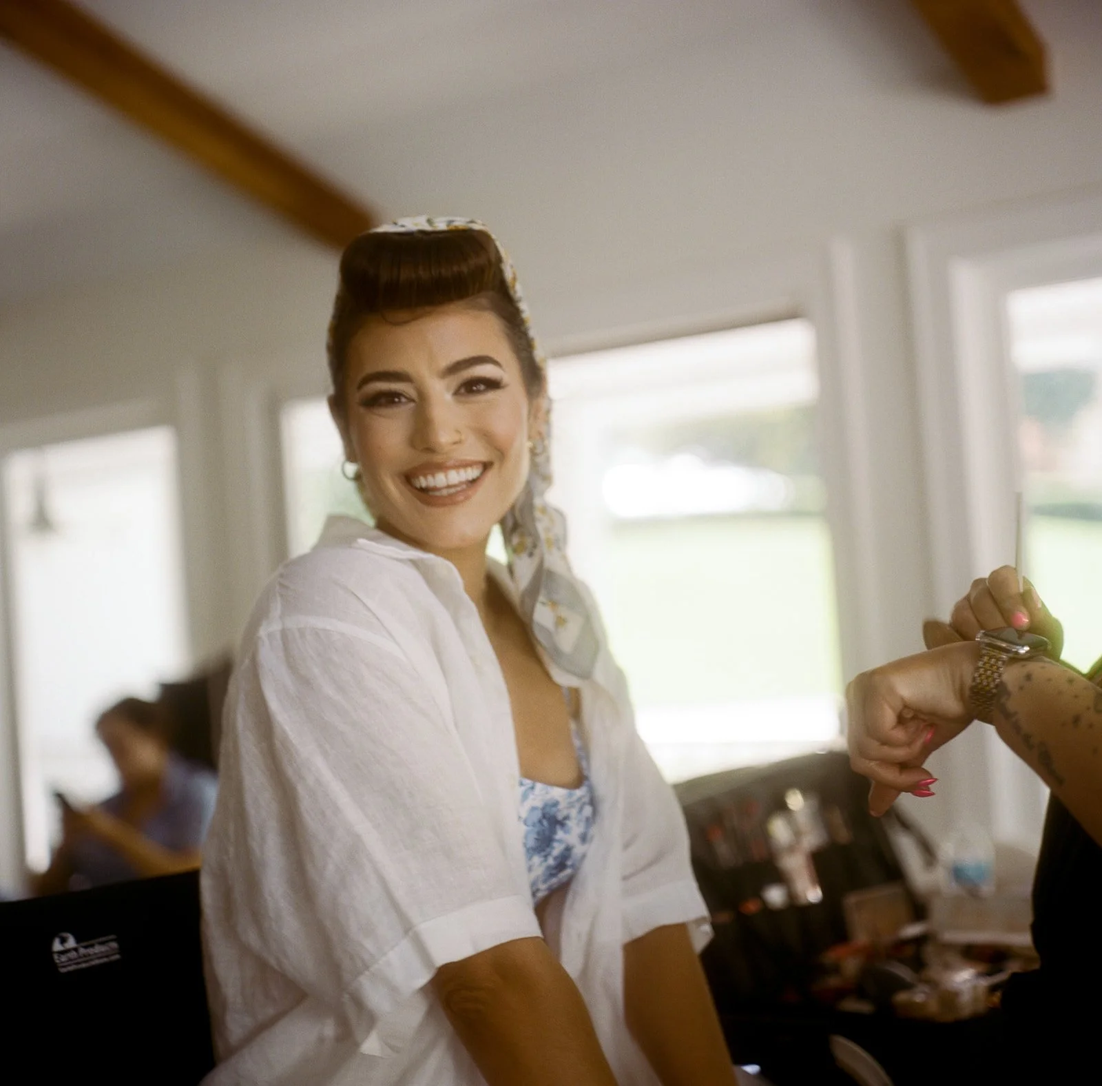 A smiling woman with vintage hairstyle and makeup, wearing a white shirt, looks at the camera while another person's arm checks their wristwatch in a bright indoor setting. shot on film on the rolleiflex in a documentary style. 