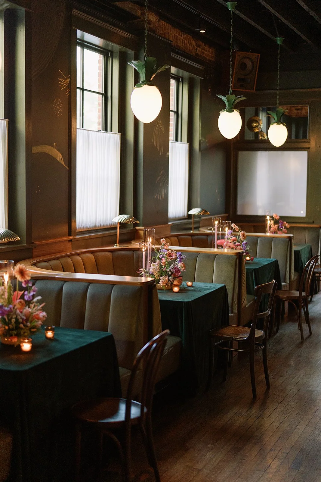 Elegant restaurant with green velvet booths, dark wooden chairs, flower centerpieces, candlelit tables, hanging pendant lights, and windows with sheer curtains.