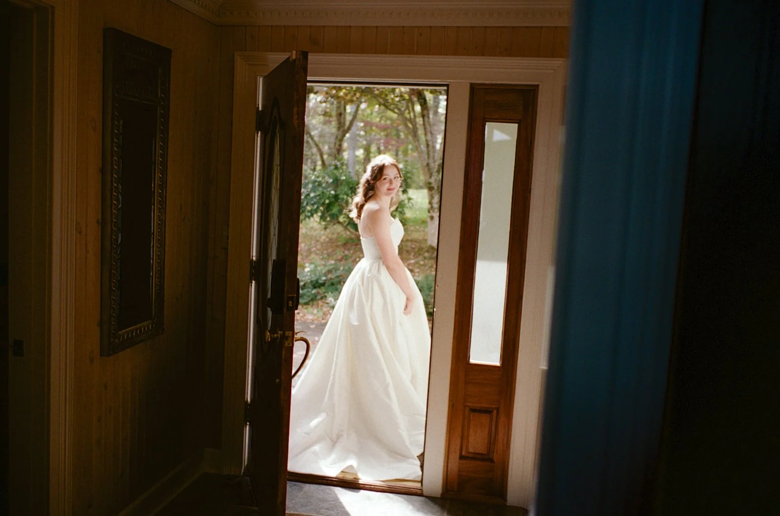 A woman in a white wedding dress standing outside a house, seen through a partially open front door.