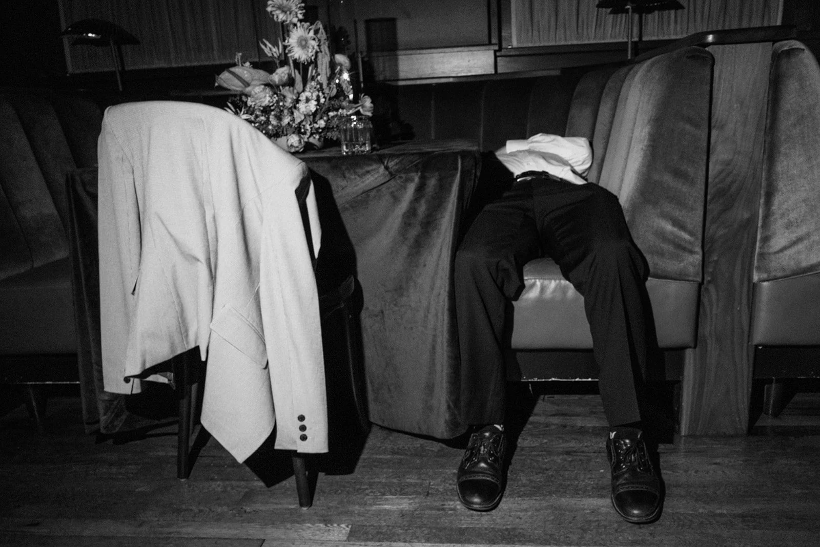 A black and white photo of a restaurant or cafe table with a floral centerpiece, a jacket draped over a chair, a man in formal attire with a white shirt and dark pants slumped over the booth with his head on the table, and his legs extending out onto