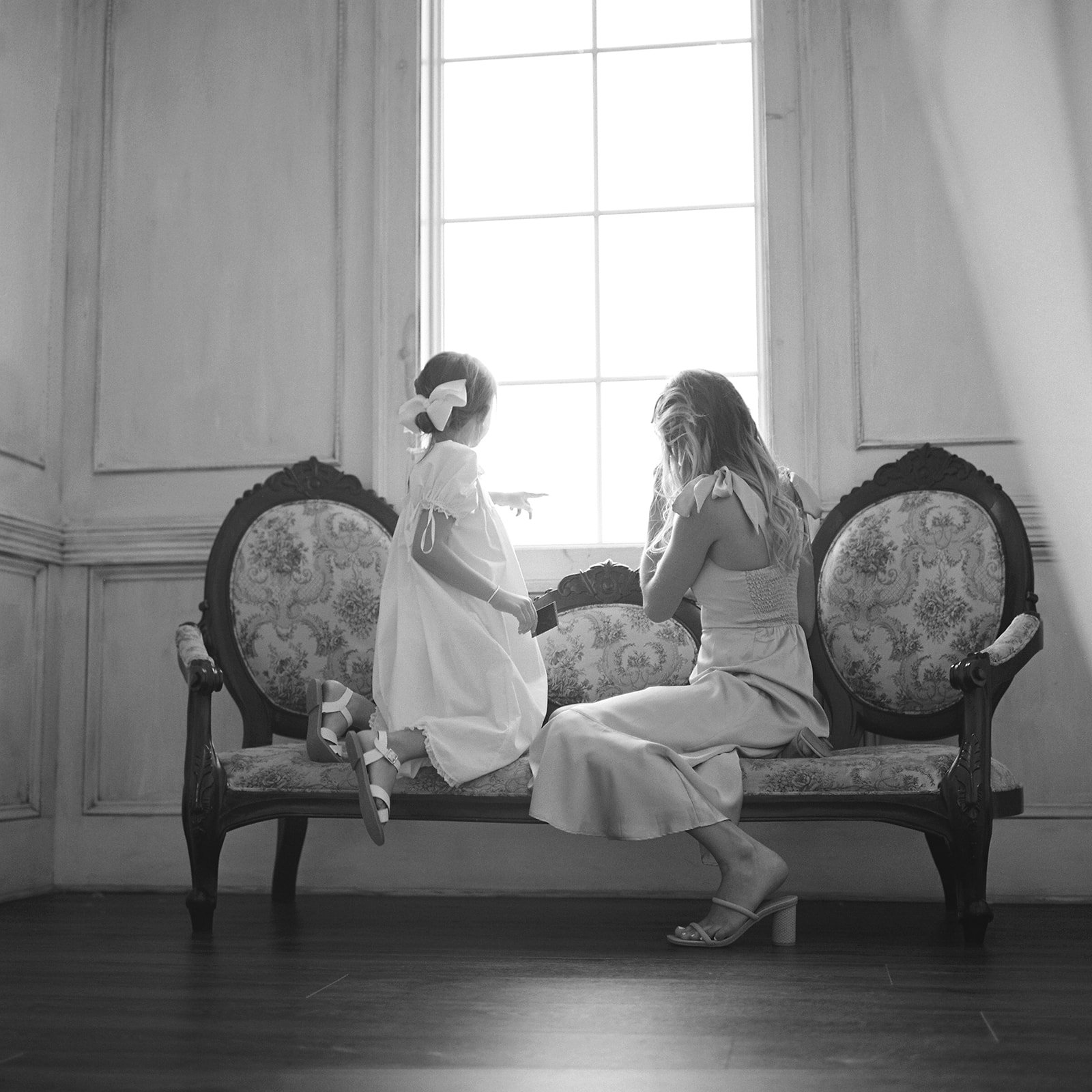 A woman and a young girl are sitting and kneeling on a vintage sofa by a large window, engaging in a conversation or play in a well-lit room.