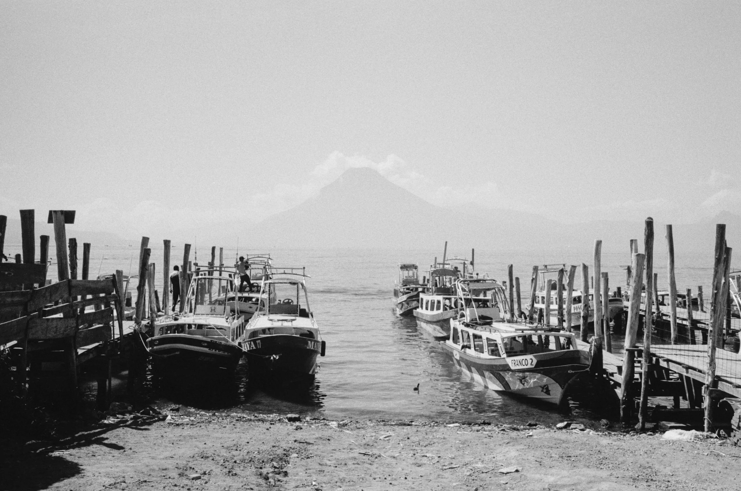 Boats docked at a harbor with a mountain in the background, in black and white.