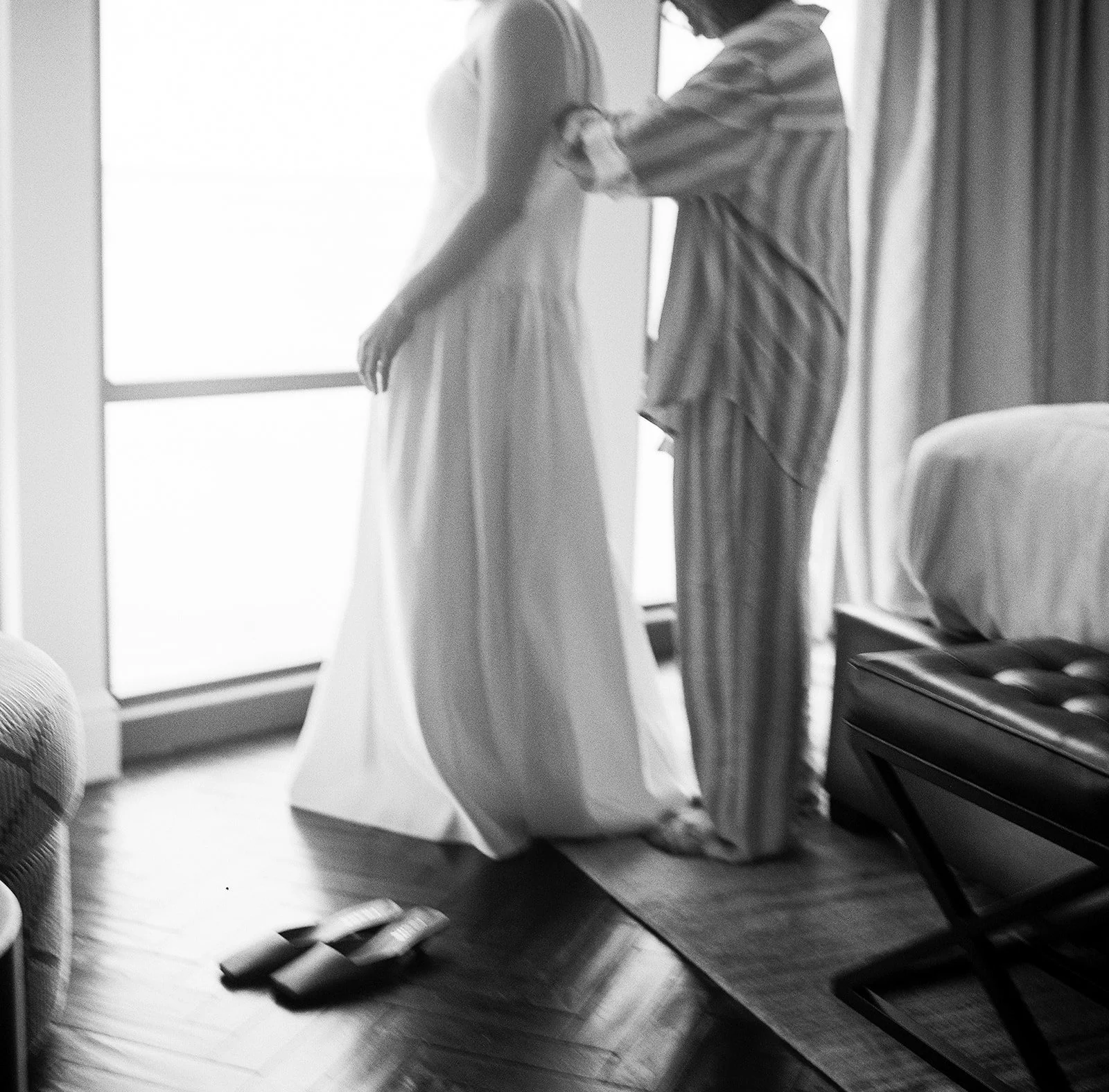A bride in a wedding dress standing near a window with a person in pajamas helping her with her dress.