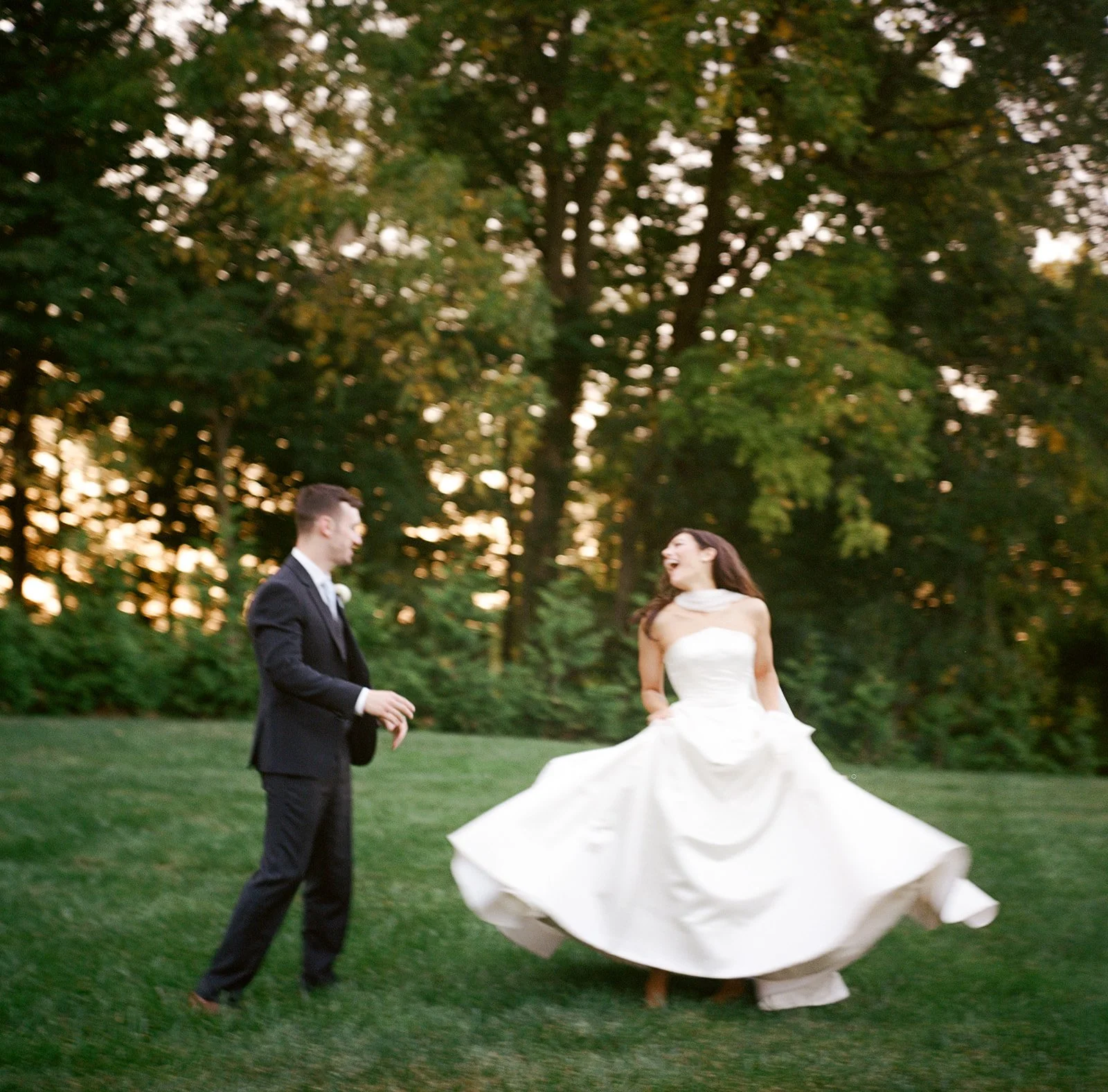 A bride in a white wedding dress and a groom in a black suit dancing outdoors on a grassy area with trees in the background during sunset.
