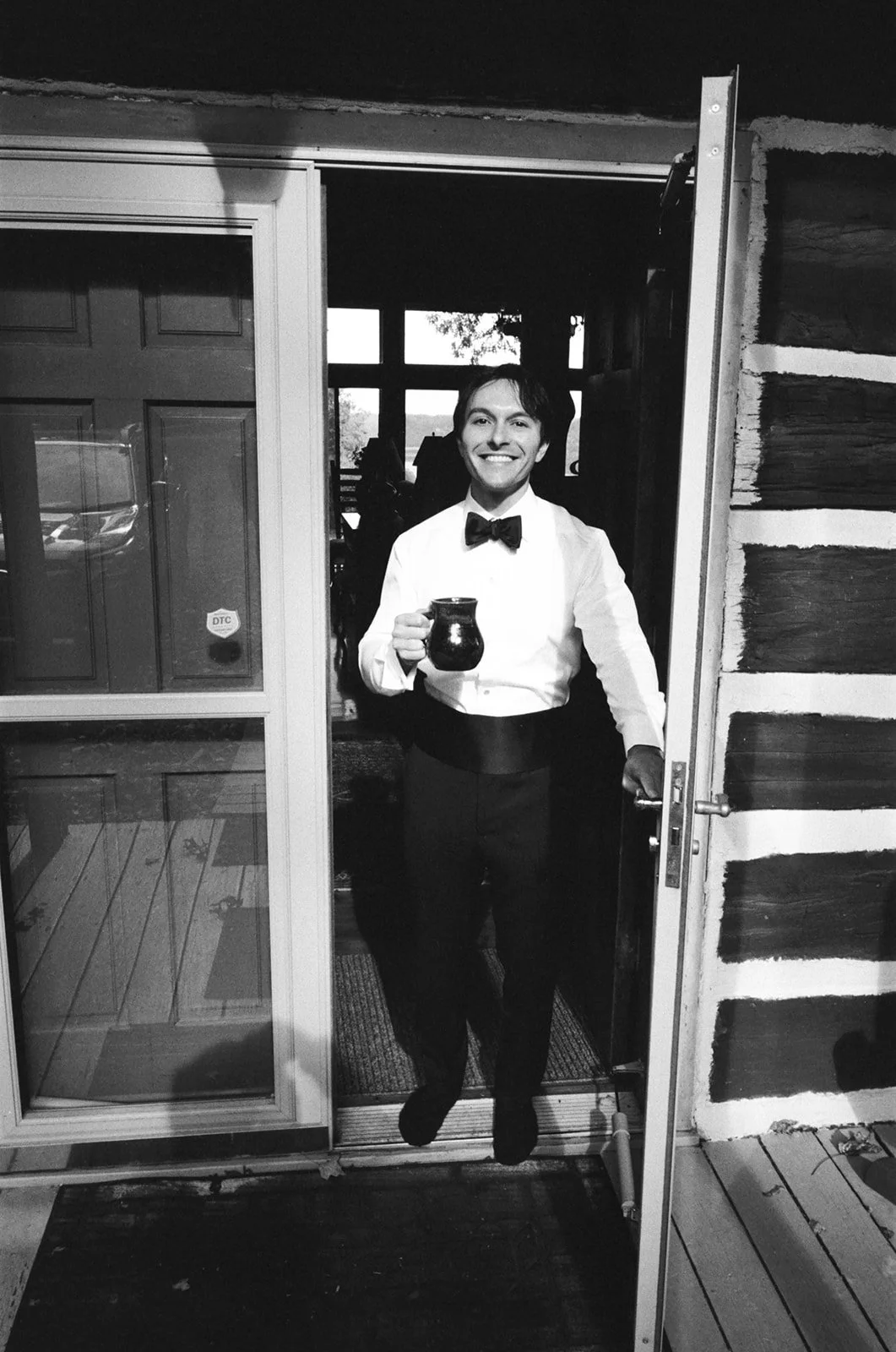 A smiling man in tuxedo and bowtie holding a coffee mug, standing at an open door of a cabin-like structure with wooden walls.