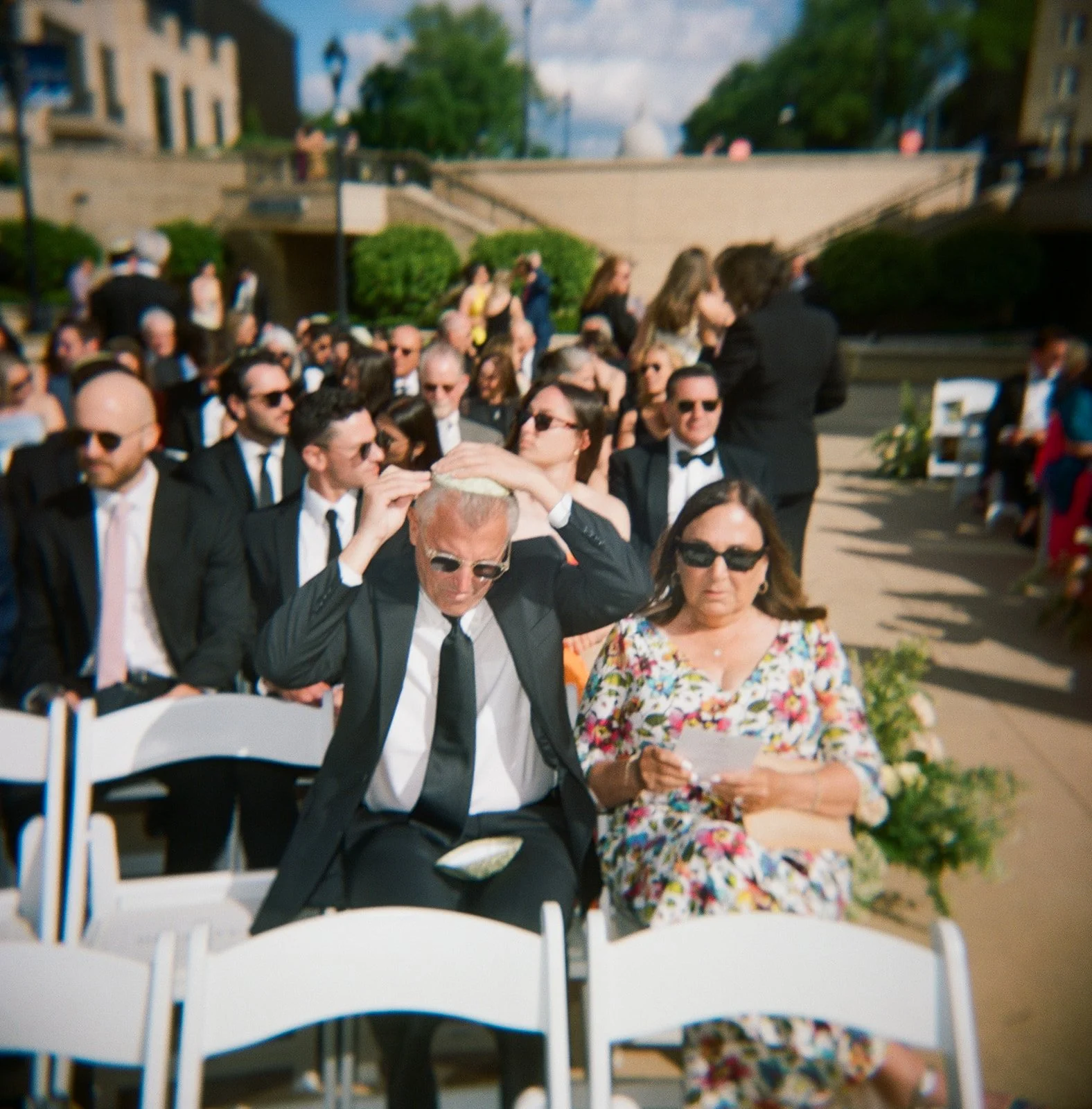 People attending an outdoor wedding ceremony, with some adjusting the officiant's head covering.
