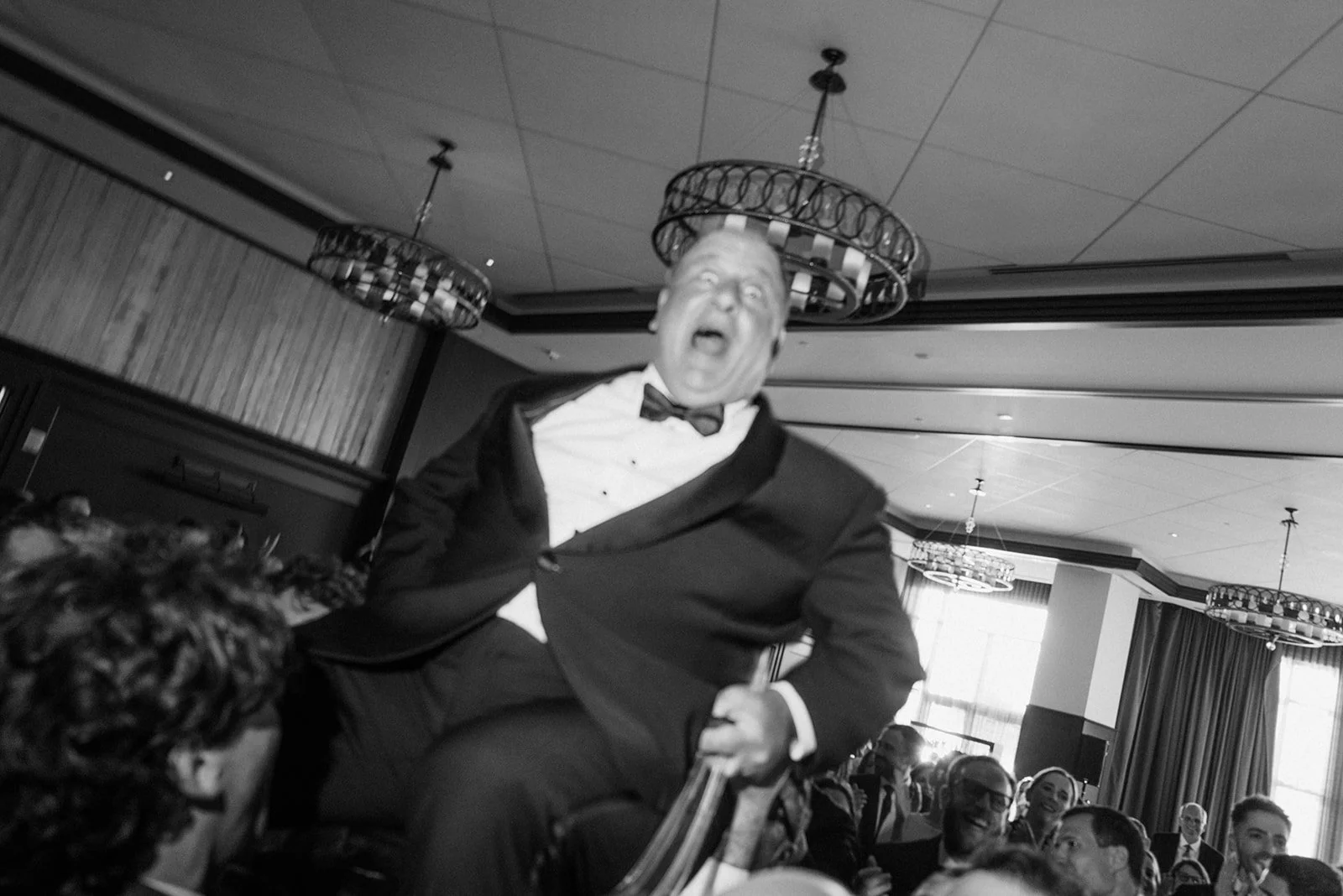 A man in formal attire, including a tuxedo and bow tie, sitting on a chair and appearing to be laughing or shouting, with people seated around him at an event or banquet in a decorated room.