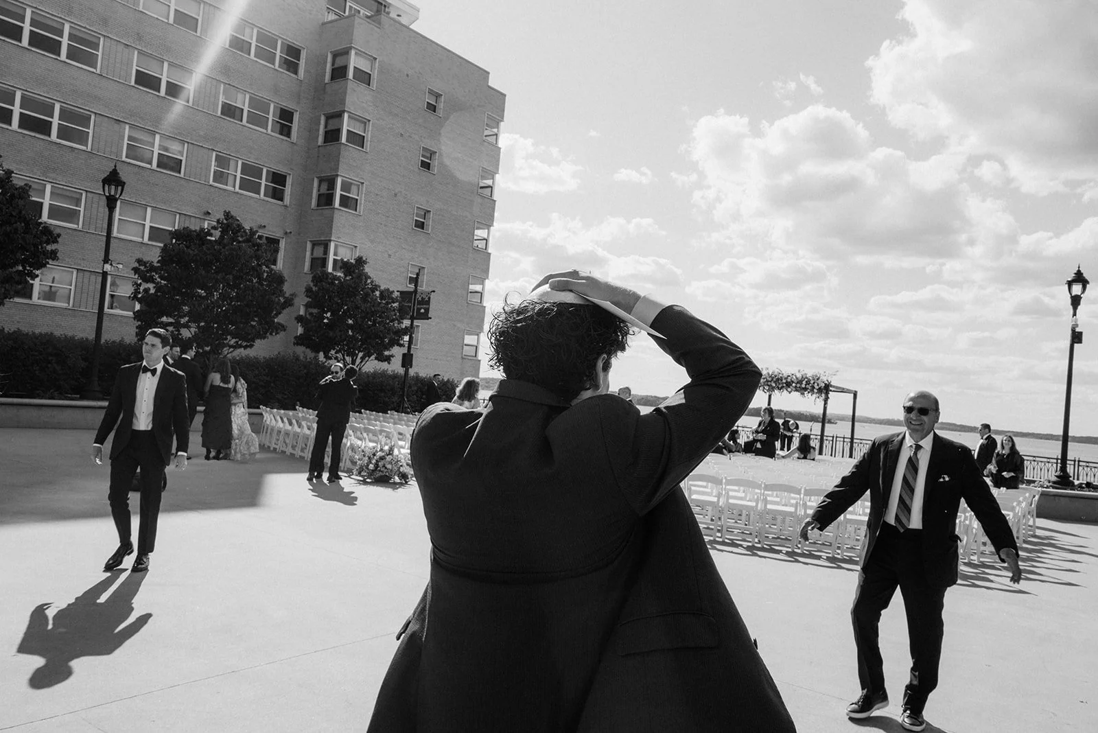 Couple dancing and smiling at an outdoor wedding reception near a river, with guests and a modern building in the background.