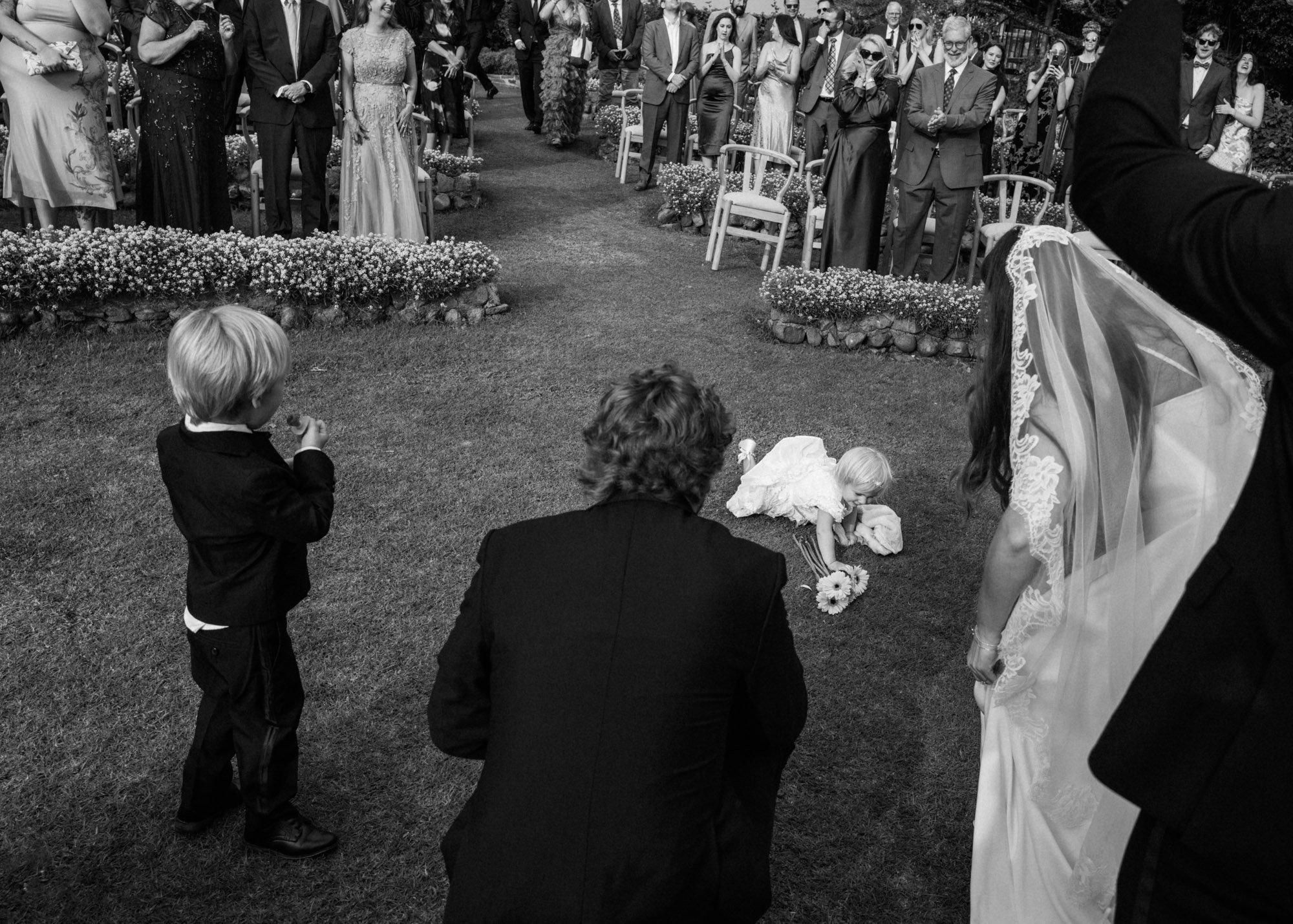 A black and white photo of a wedding ceremony outdoors. A bride is bending down to adjust her shoe, while a young flower girl lies on the ground with a bouquet nearby. Two young boys and several adults, dressed in formal attire, are watching in the b