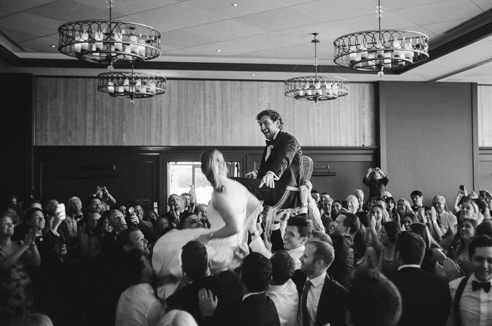 A black and white photo of a wedding celebration showing a groom riding on a chair, being lifted by friends, with a woman, likely the bride, sitting on his lap. The guests are smiling, cheering, and capturing the moment with their phones in a large b