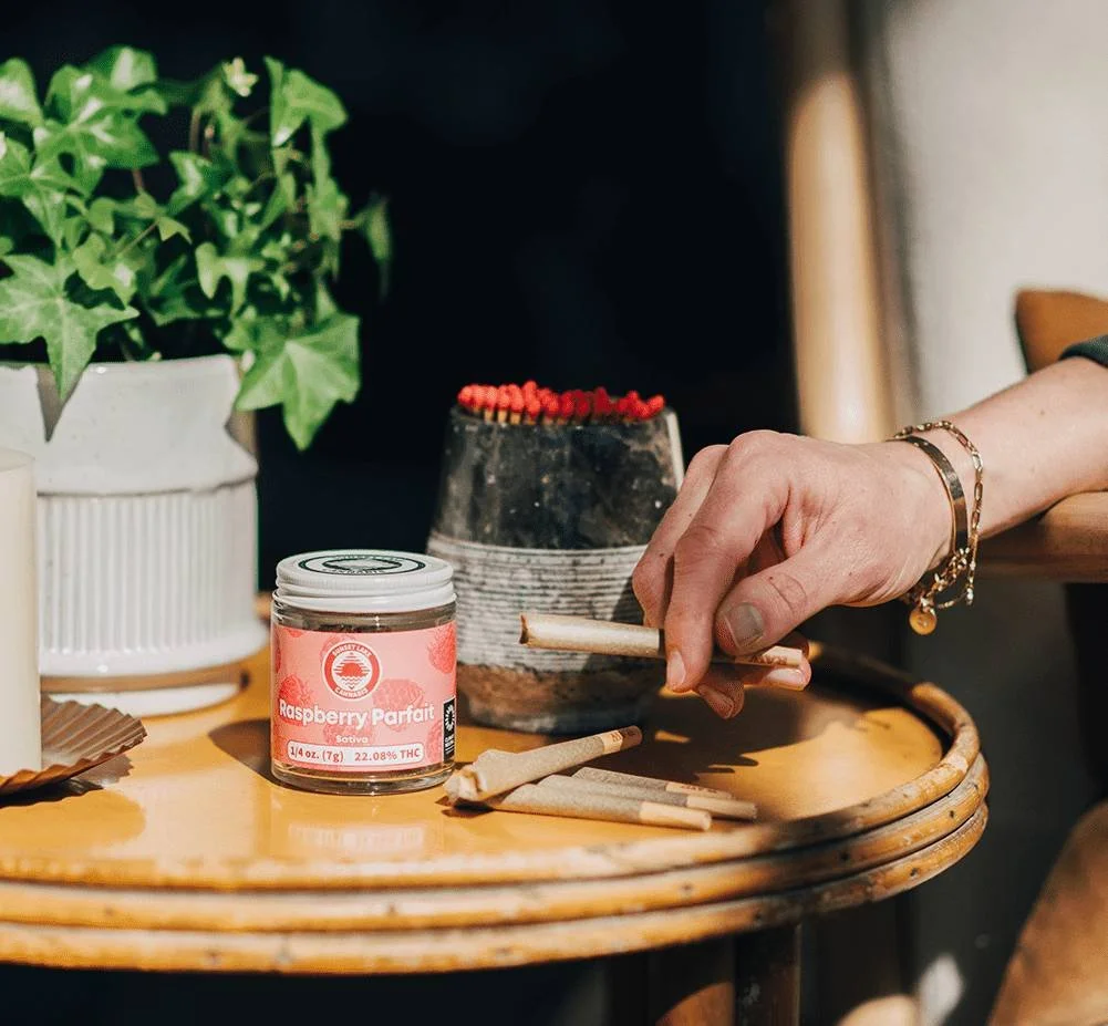 Side table with jar of Sunset Lake Cannabis Raspberry Parfait and a pile of joints and stone jar of matches. A hand is picking up a joint.