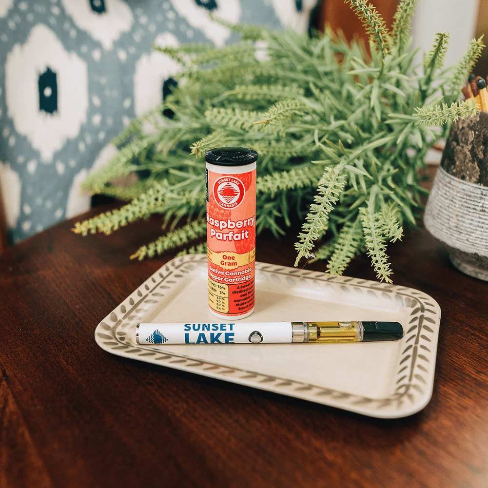 Photo of a Sunset Lake Cannabis Raspberry Parfait vape cartridge and battery on a decorative tray in front of a house plant.