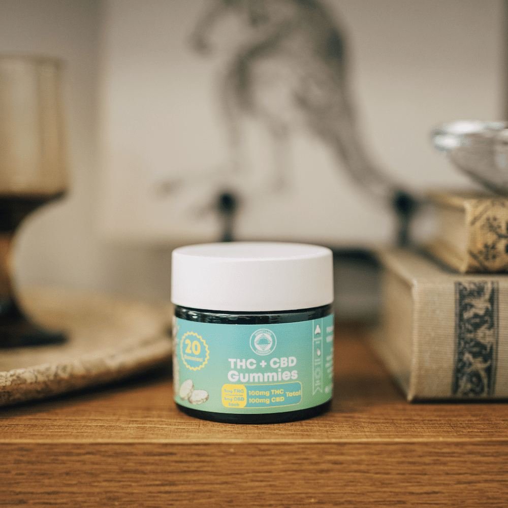 Photo of a jar of Sunset Lake Cannabis THC + CBD pineapple-flavored gummies on a bookshelf with some nicknacks. Buy Sunset Lake Cannabis THC + CBD gummies at a Vermont dispensary near you.