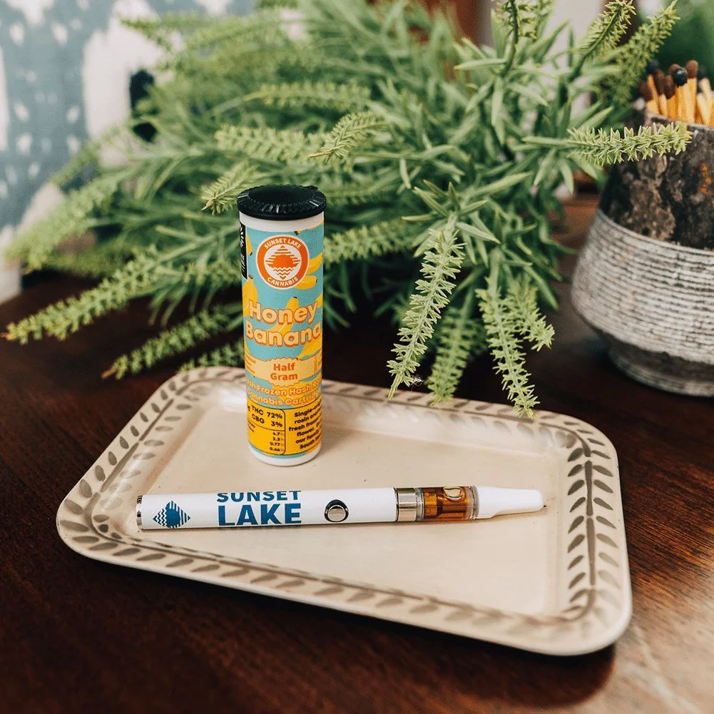Photo of a Sunset Lake Cannabis Honey Banana live hash rosin vape cart and battery on a decorative tray in front of a house plant and a stone jar of matches.