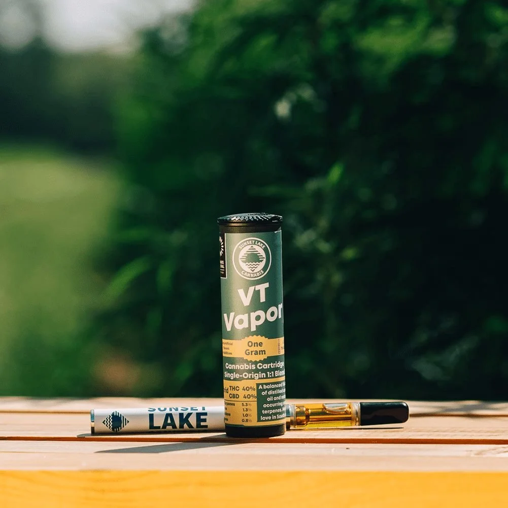 Photo of a Sunset Lake Cannabis VT Vapor Hybrid 1-to-1 THC + CBD vape cartridge on a wooden box in a cannabis field.