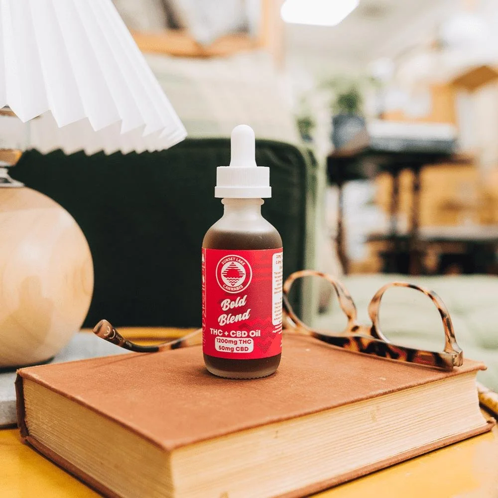 Bedside table with a dropper bottle of Sunset Lake Cannabis Bold Blend THC Tincture on top of a book next to a lamp and a pair of reading glasses.
