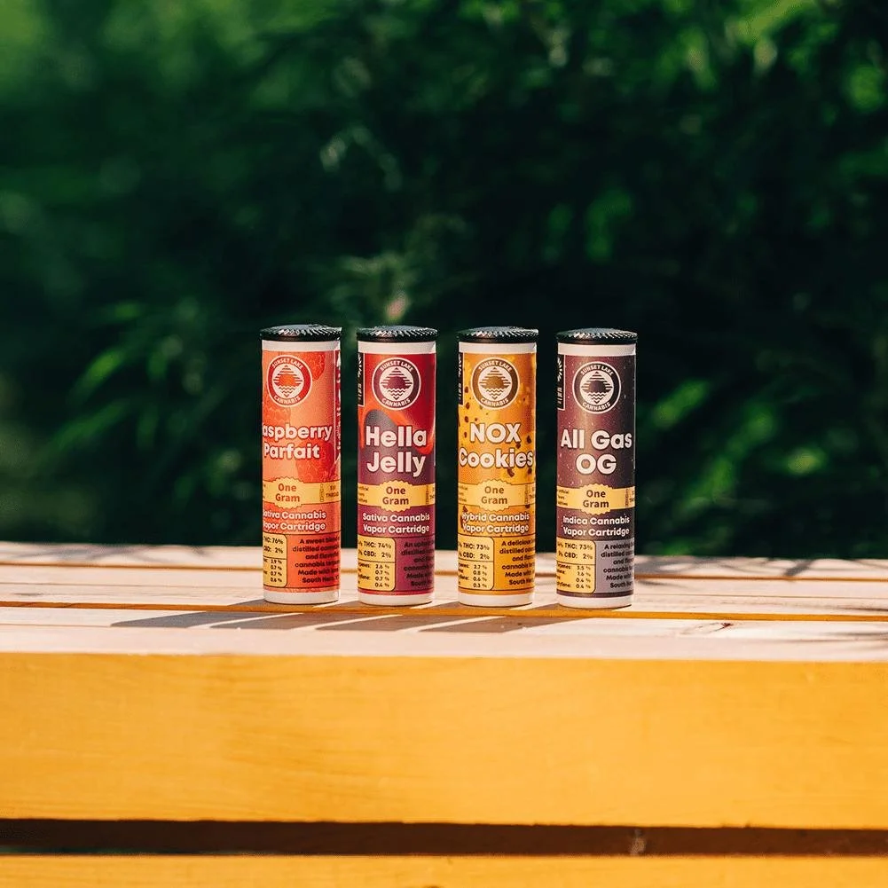 Sunset Lake Cannabis vape carts on a wooden crate in front of a cannabis plant. From left to right: Raspberry Parfait, Hella Jelly, NOX Cookies, All Gas OG.