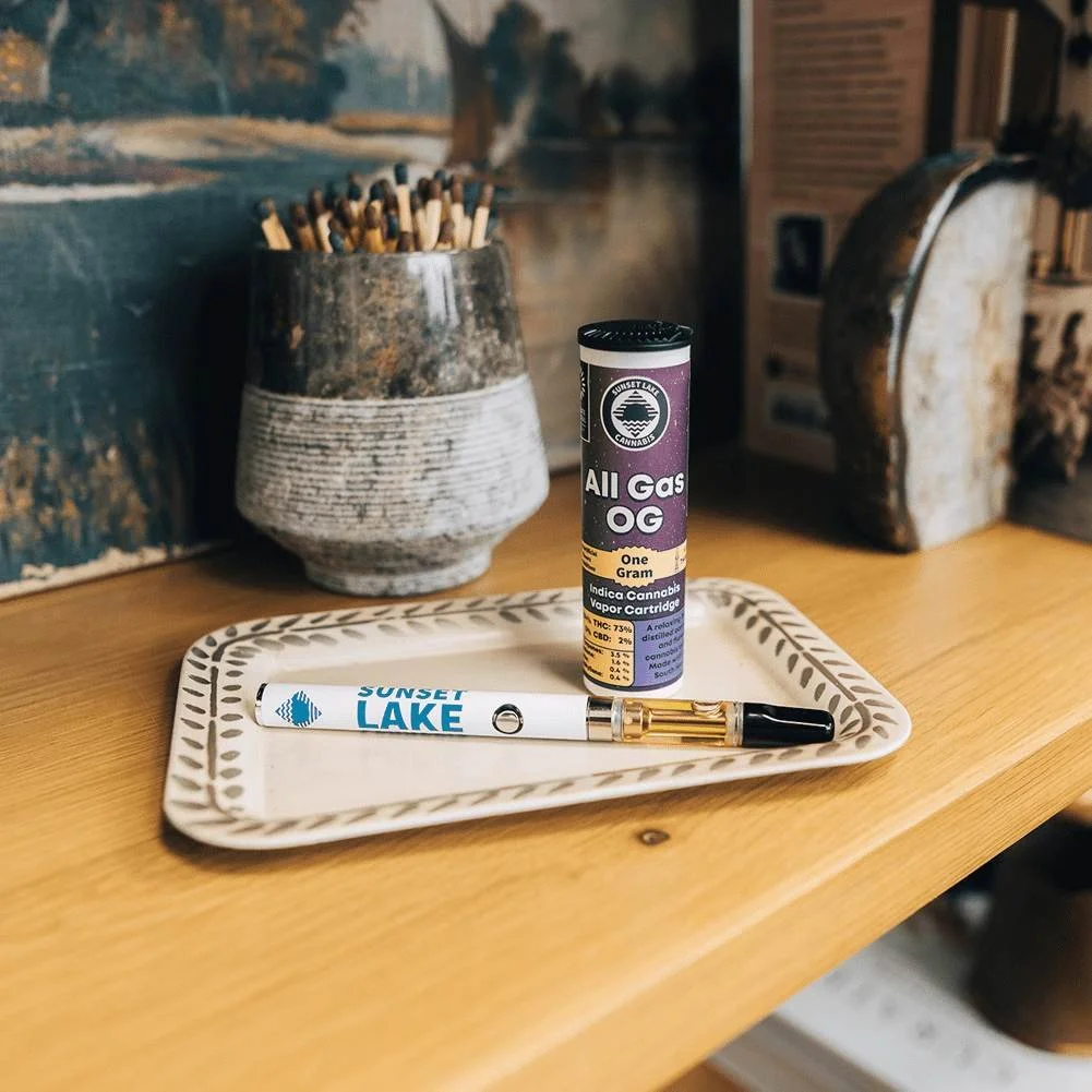 Photo of a Sunset Lake Cannabis All Gas OG vape cart and battery sitting on a bookshelf with a painting, books, and a stone jar of matches.