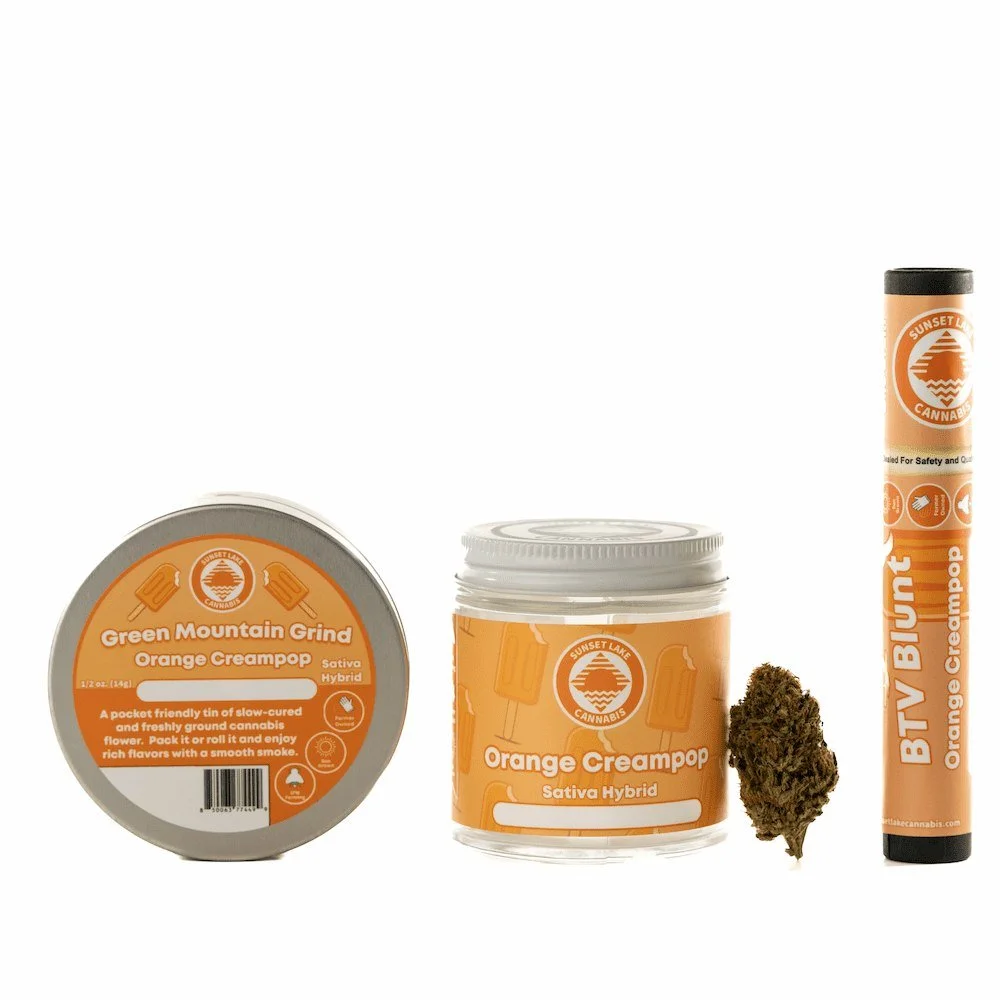 Photo of a group of Sunset Lake Cannabis Orange Creampop stain products including a tin of Green Mountain Grind, a quarter ounce jar of hand-trimmed flower, a loose bud, and a BTV Blunt.