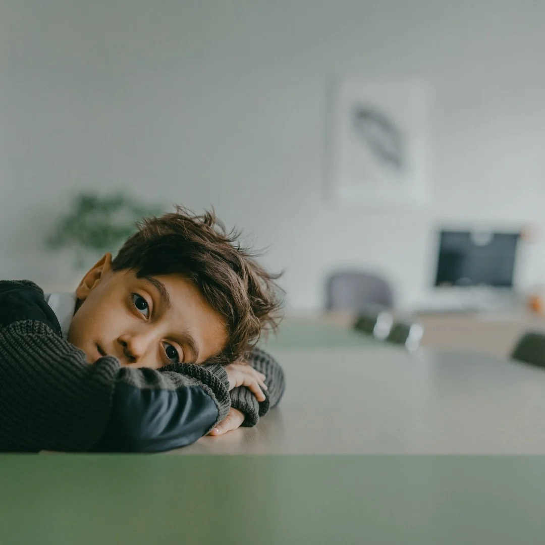 A young boy rests his head on a table, looking thoughtfully into the distance in a softly lit classroom.