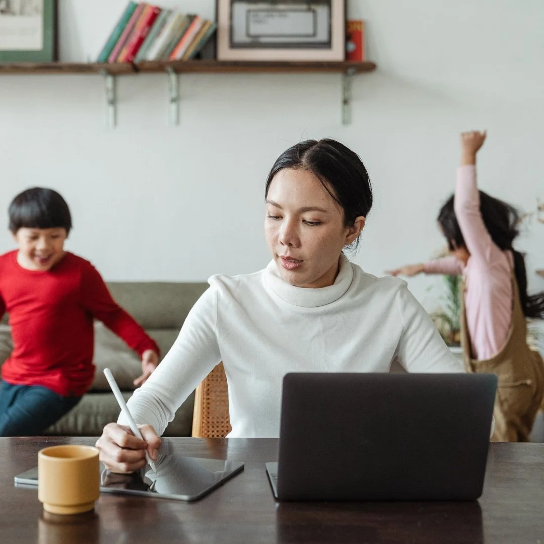 Woman working at a desk with a laptop and tablet while two children play energetically in the background.