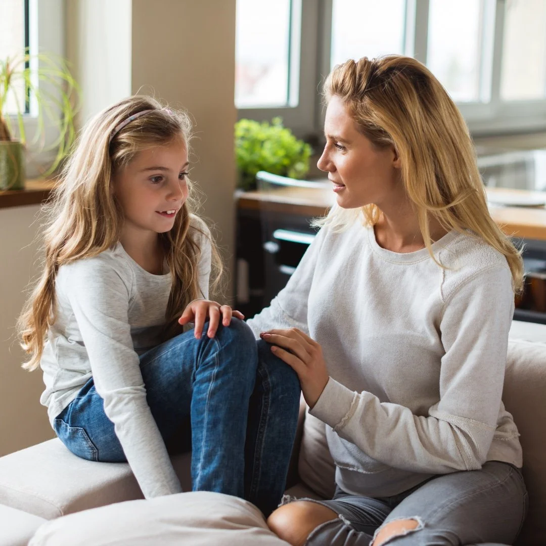A woman and a young girl sit on a couch, facing each other and talking in a bright, cozy living room.