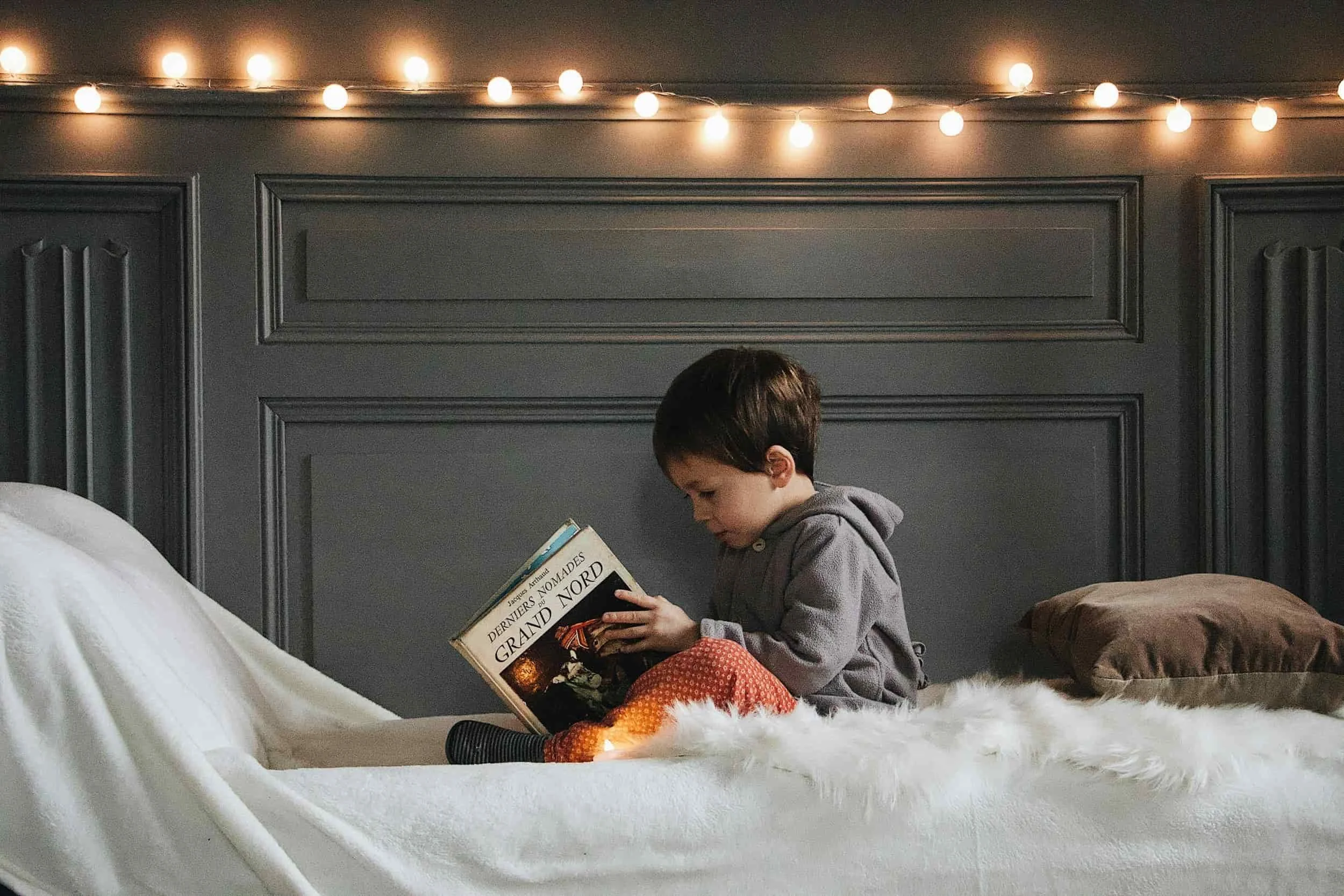 Young child sitting on a bed reading a book, with warm string lights hanging on a gray wall in the background.
